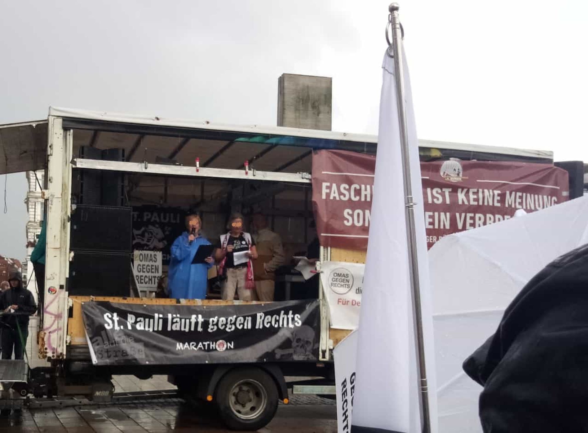 Blick auf den Lautsprecherwagen. Zwei ältere Frauen halten Klemmbretter mit Zetteln und sprechen in Mikrofone. Unter ihnen hängt ein Banner des FC St. Pauli "St. Pauli läuft gegen Rechts / Marathon", wobei das O in Marathon aus dem St. Pauli Logo besteht.