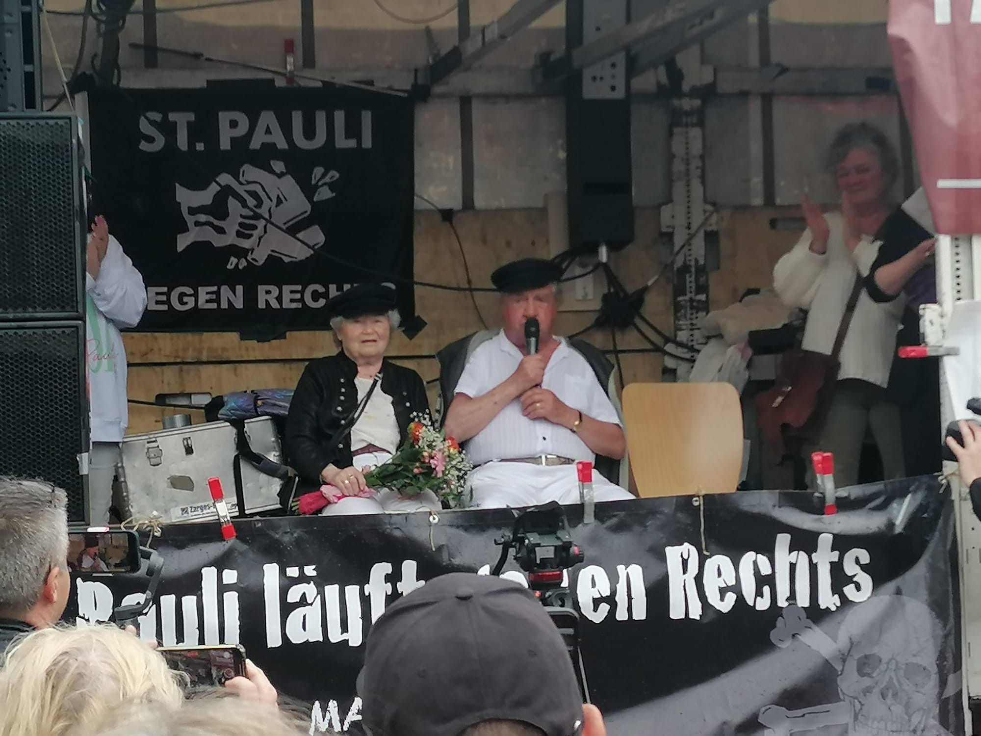 Der Lautsprecherwagen des FC St. Pauli. An der hinteren Wand hängt eine schwarze Fahne mit weißer Aufschrift: "St. Pauli gegen Rechts", Mittig das Bild einer Faust, die ein Hakenkreuz zerschlägt. 
In der Mitte des Wagens sitzt Ivar Butterfaß mit seiner Frau. Sie hält einen Blumenstrauß, er spricht in ein Mikrofon.