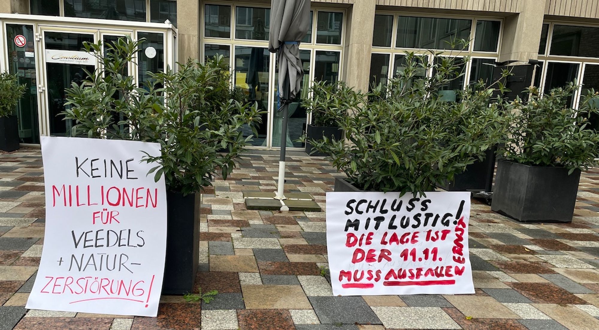 Zwei Demonstrationsplakate vor dem Kölner Rathaus. Links: "Keine Millionen für Veedels+Natur-Zerstörung". Rechts: "Schluss mit lustig! Die Lage ist: Der 11.11. muss ausfallen"