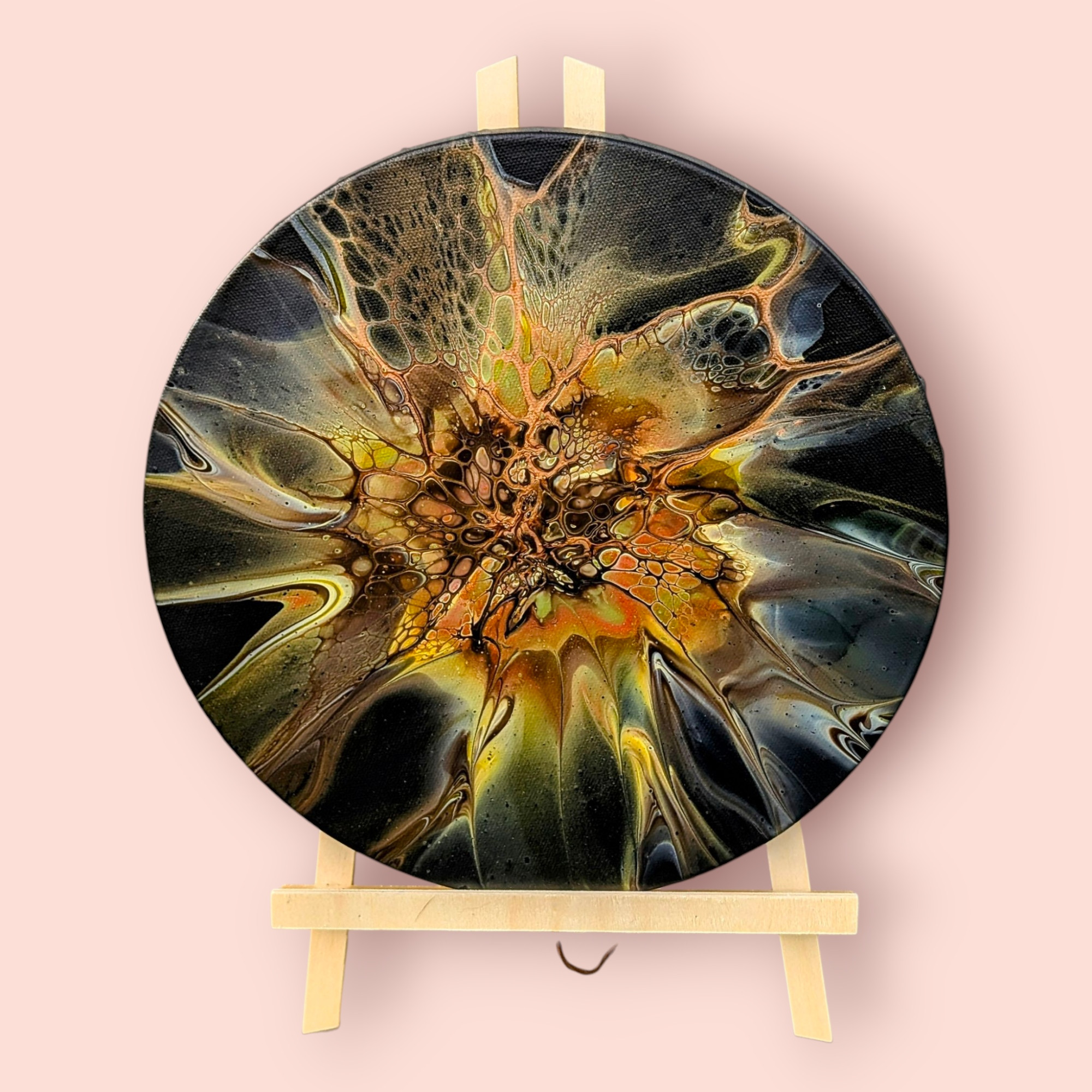 A fluid art bloom displayed on a wooden easel over a very soft peachy background; the background of the painting itself is black, the main colors are primarily shades of yellow along with a hint of deep orange and olive green, and the cell activators are Van Dyke brown and old gold.