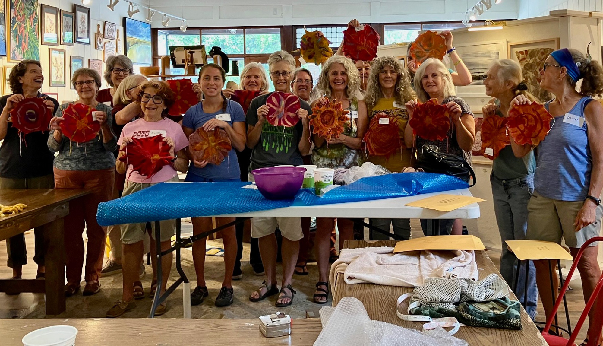 A group of about 15 elders stands in the gallery workshop space behind a table with crafting supplies. Each person holds a felted sun in orange, gold, red, or purple.