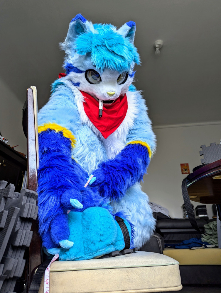 Linko assembling his fursuit sandals. His paw is pose don a chair, and he measures the length of a strap.