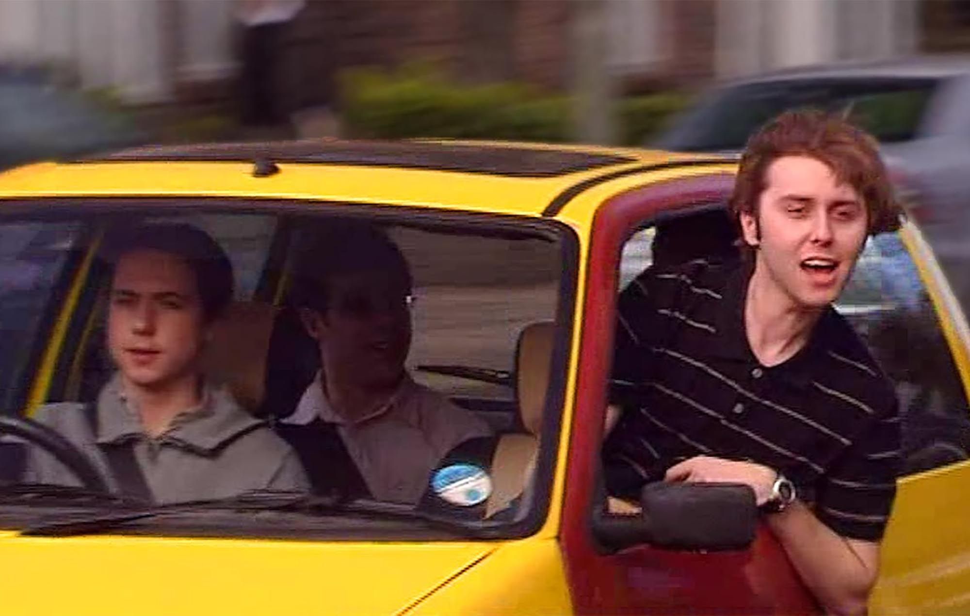 still of 'bus stop wankers' from the television show the Inbetweeners.
A right-hand drive yellow car with a brown passenger side door is driving from right to left. There are 4 white teenage boys inside. One of whom is leaning out of the brown-door passenger window to yell 'bus stop wankers' at some school kids out of frame