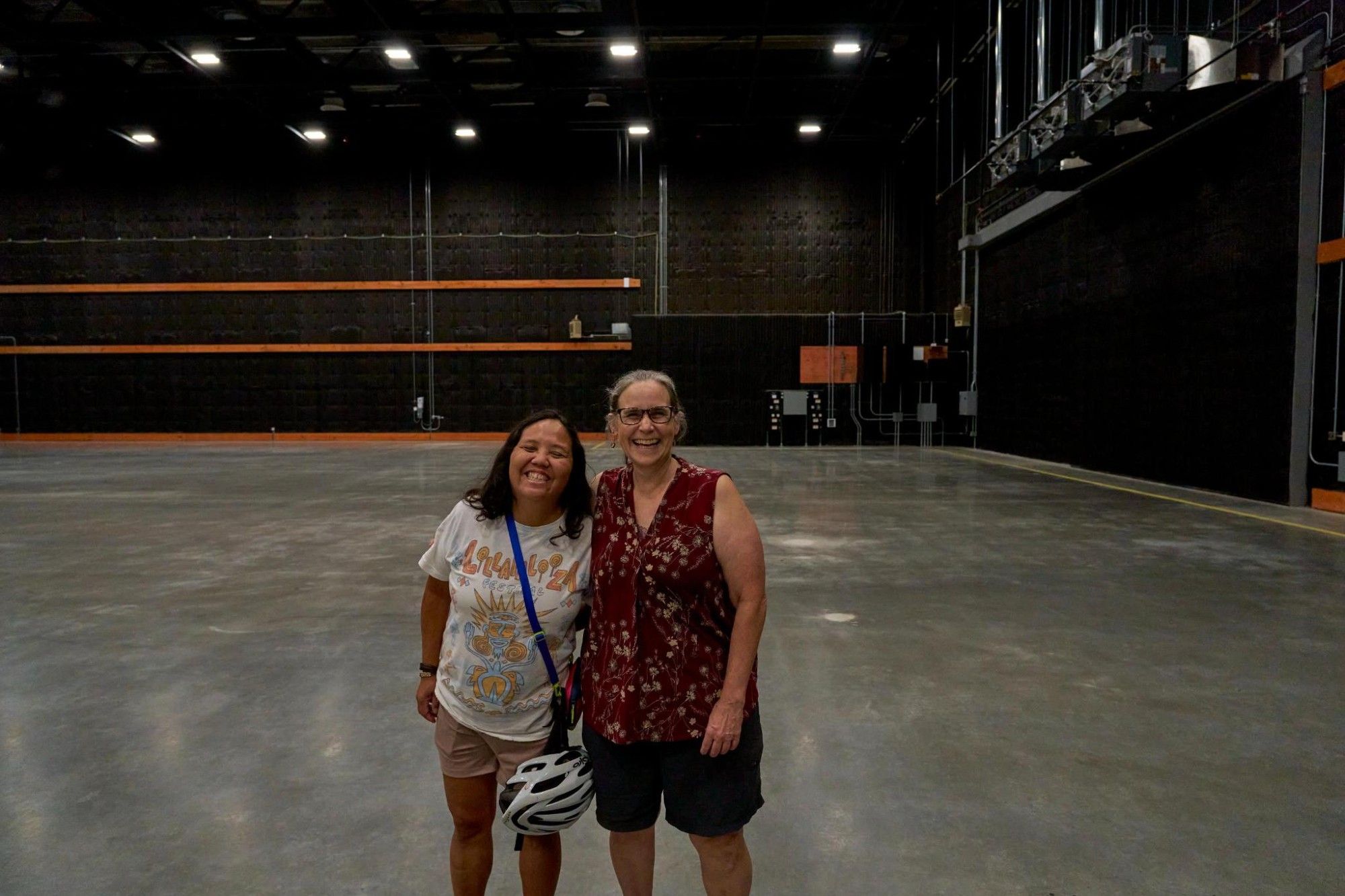 My neighbor, uber-cool kindie musician Little Miss Ann, checking out the space. Two women stand in the foreground smiling. In the background is a grey, shiny floor extending to the distant walls, which are black with orange bars.