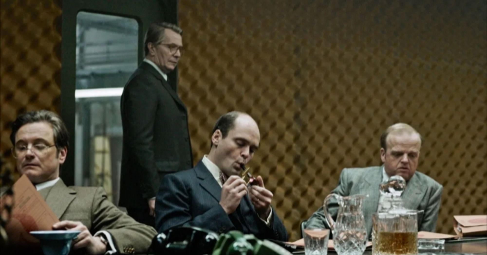 Image is of still photo of the film Tinker Tailor Soldier Spy starring Gary Oldham. In this image Smiley is walking in two briefing room where's three of his subordinates are seated.