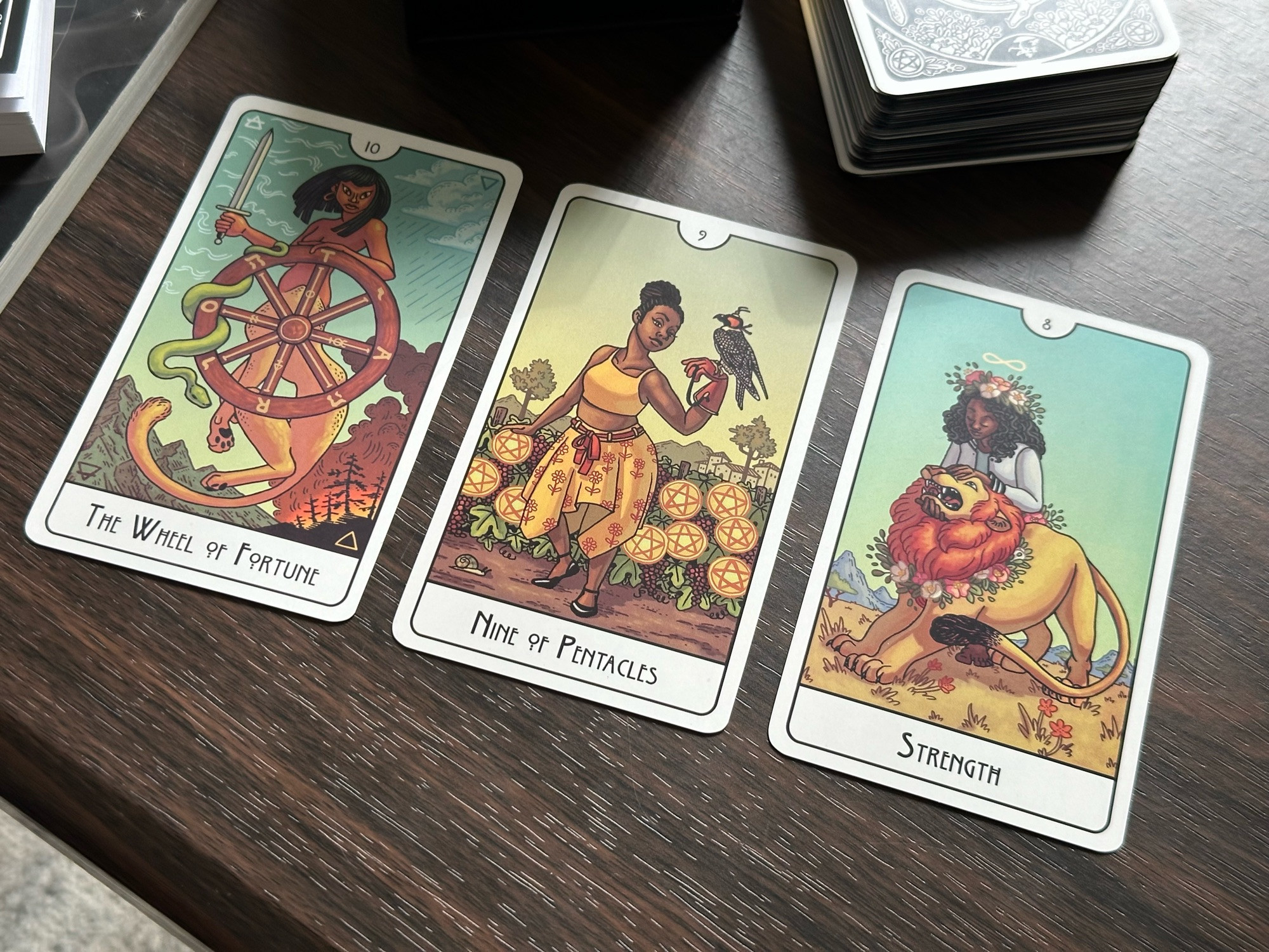 A three-card tarot spread featuring the Wheel of Fortune, Nine of Pentacles, and Strength. The deck is the This Might Hurt Tarot by Isabella Rotman. 