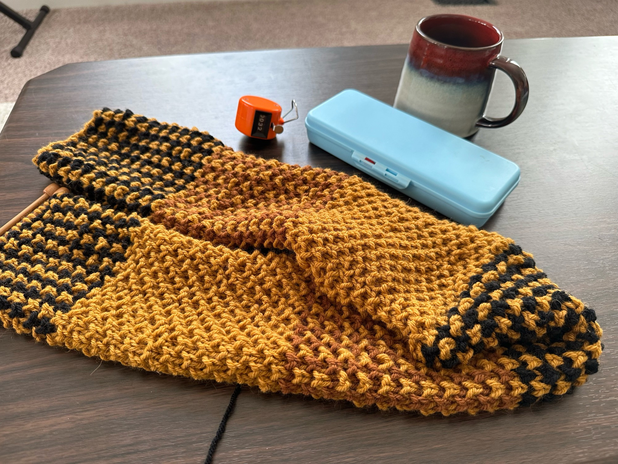 A knitting project on a brown tabletop. It features stripes of dark yellow, pale brown, and black yarn to emulate a plaid pattern. The stitch is a plush brioche stitch which gives a textured look. Nearby on the table is a knitting tool kit, an orange row counter, and a cup of coffee in a ceramic mug. 