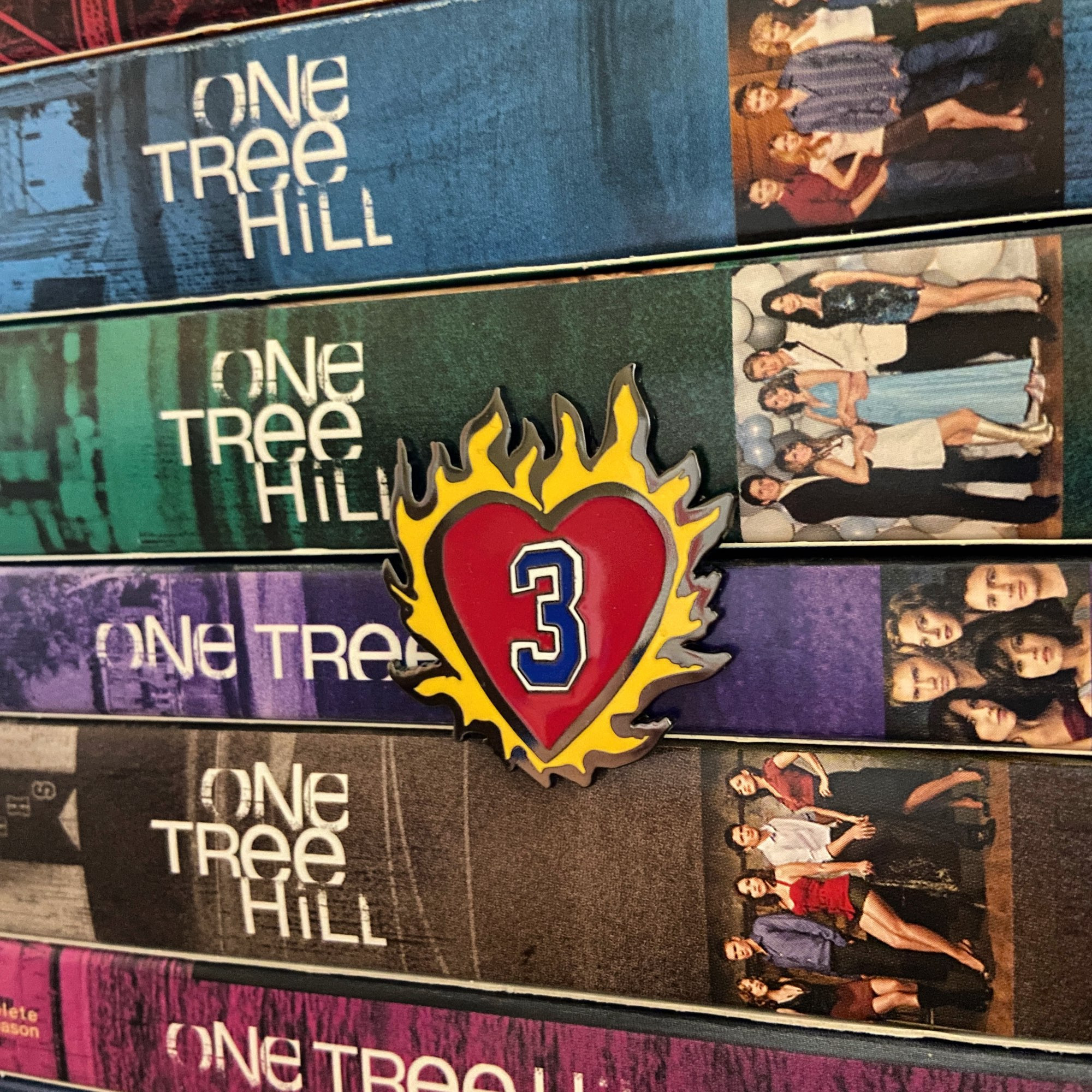 Enamel pin of the number 3 surrounded by a heart on fire. Behind the enamel pin is a stack of One Tree Hill dvds.