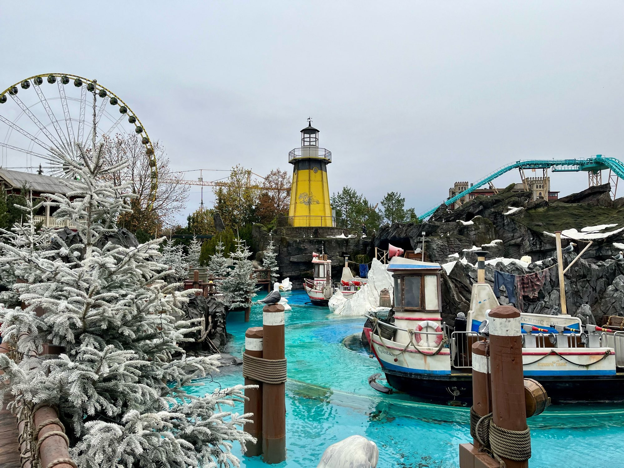 Ein Boot der Wasserbahn „Whale Adventures“  fährt durch das Bild. Seitlich stehen weiße Tannenbäume, im Hintergrund ein gelber Leuchtturm, das Riesenrad und eine andere Wasserbahn in türkis