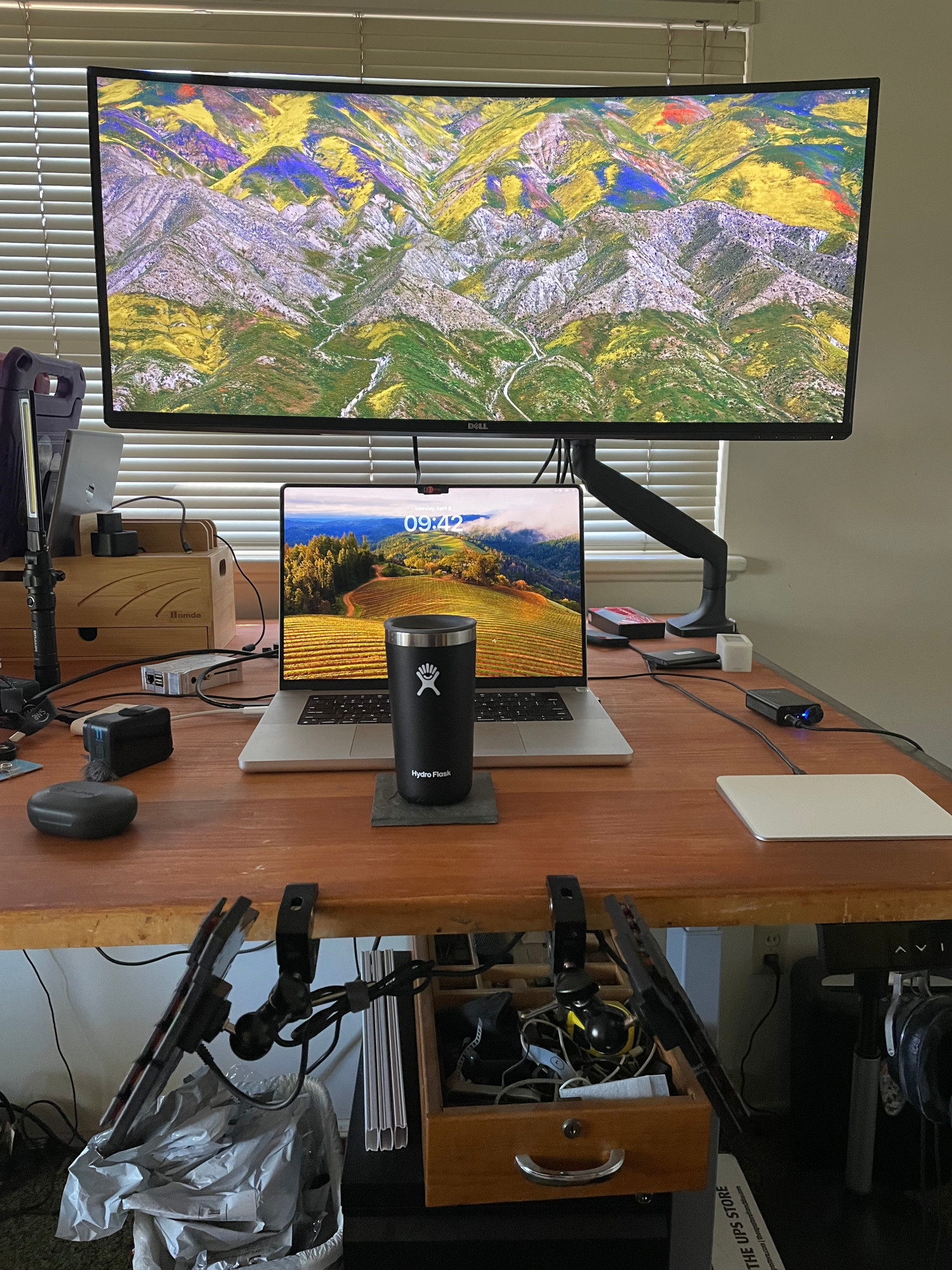 A desktop with a laptop, a widescreen monitor and a ZSA Voyager split ergonomic keyboard mounted to the edge of the desktop on small articulating tripod mount clamps.