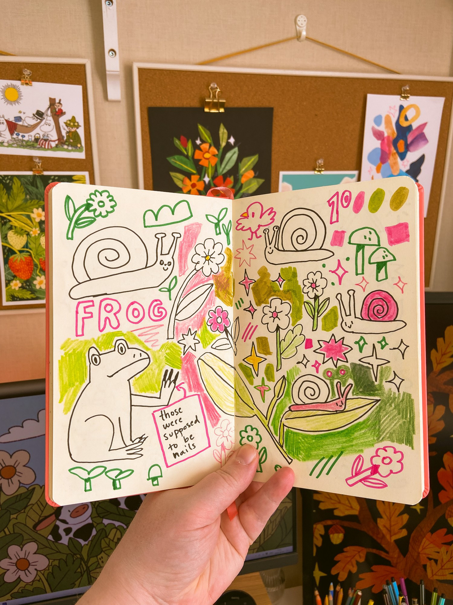 A photo of my hand holding up my sketchbook in front of my desk setup. Inside the sketchbook are lots of silly doodles, scribbles and sparkles: flowers, snails, a weird frog etc.