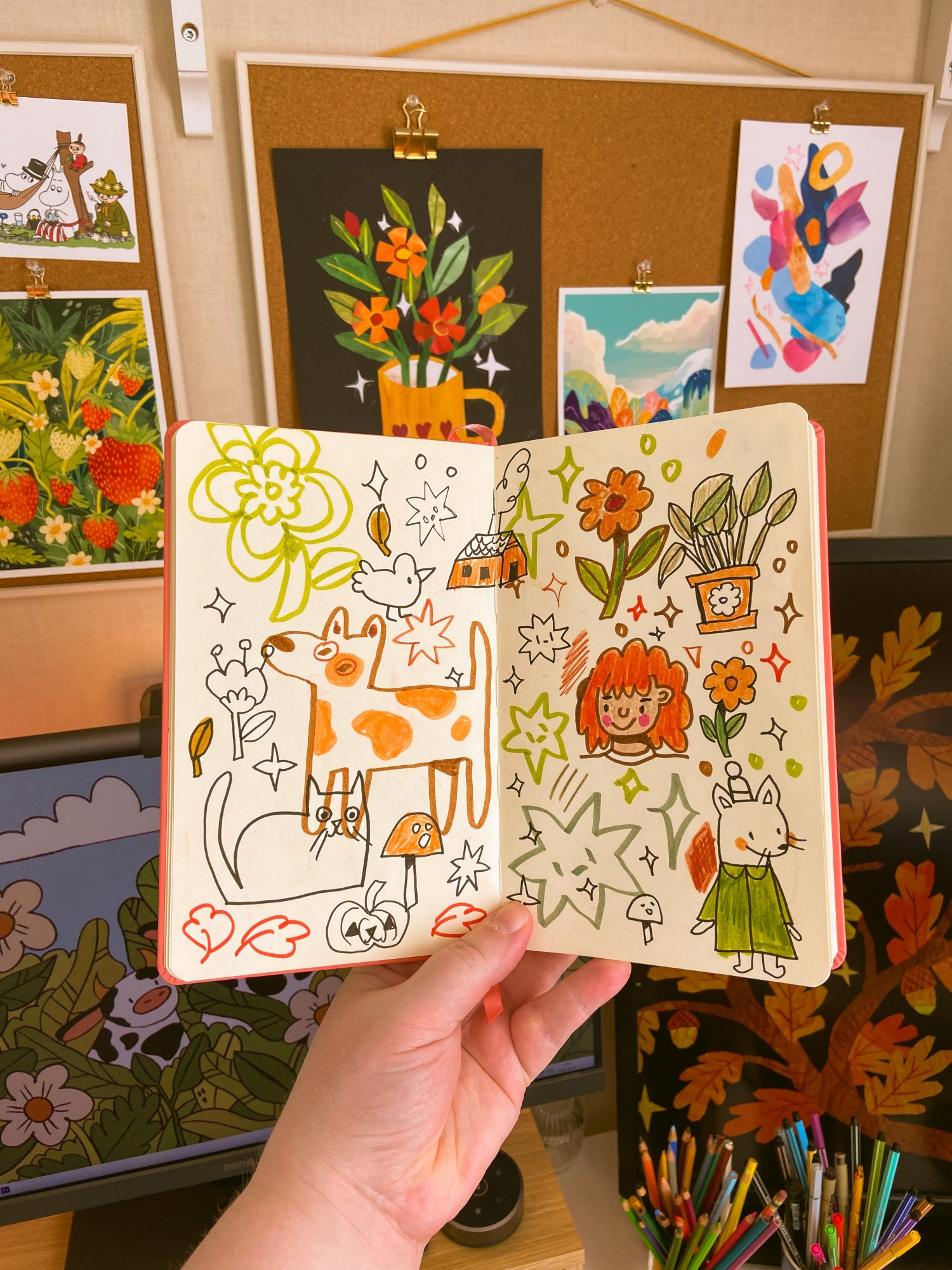 A photo of my hand holding up my sketchbook in front of my desk setup. Inside the sketchbook are lots of silly doodles, scribbles and sparkles: a dog, flowers, a girl, plants and a cat with very staring eyes.