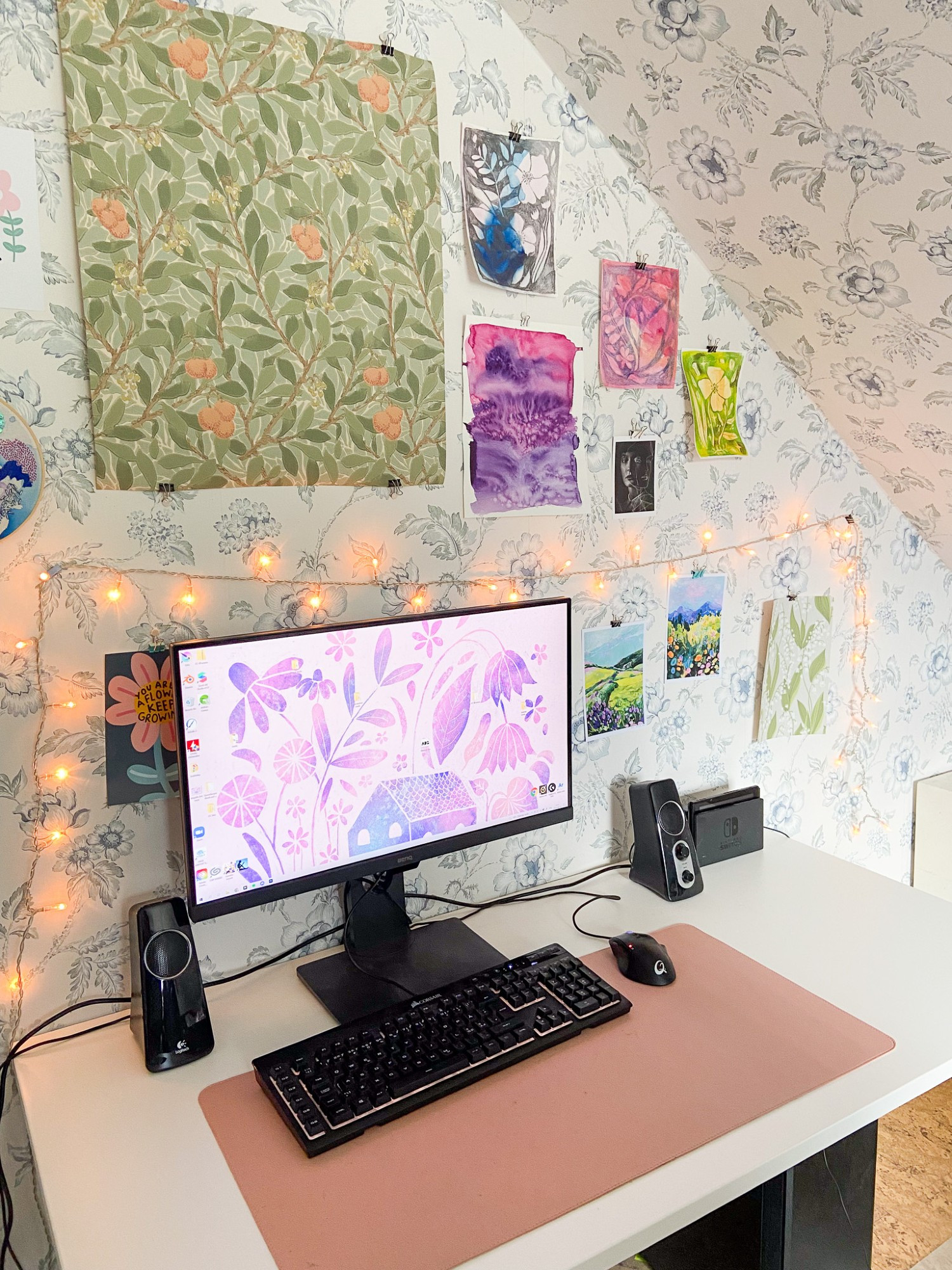 A photo of my desk space from 2021 - a white desk with black tech on it, and lots of prints on the wallpapered wall. There's a pink desk mat. 