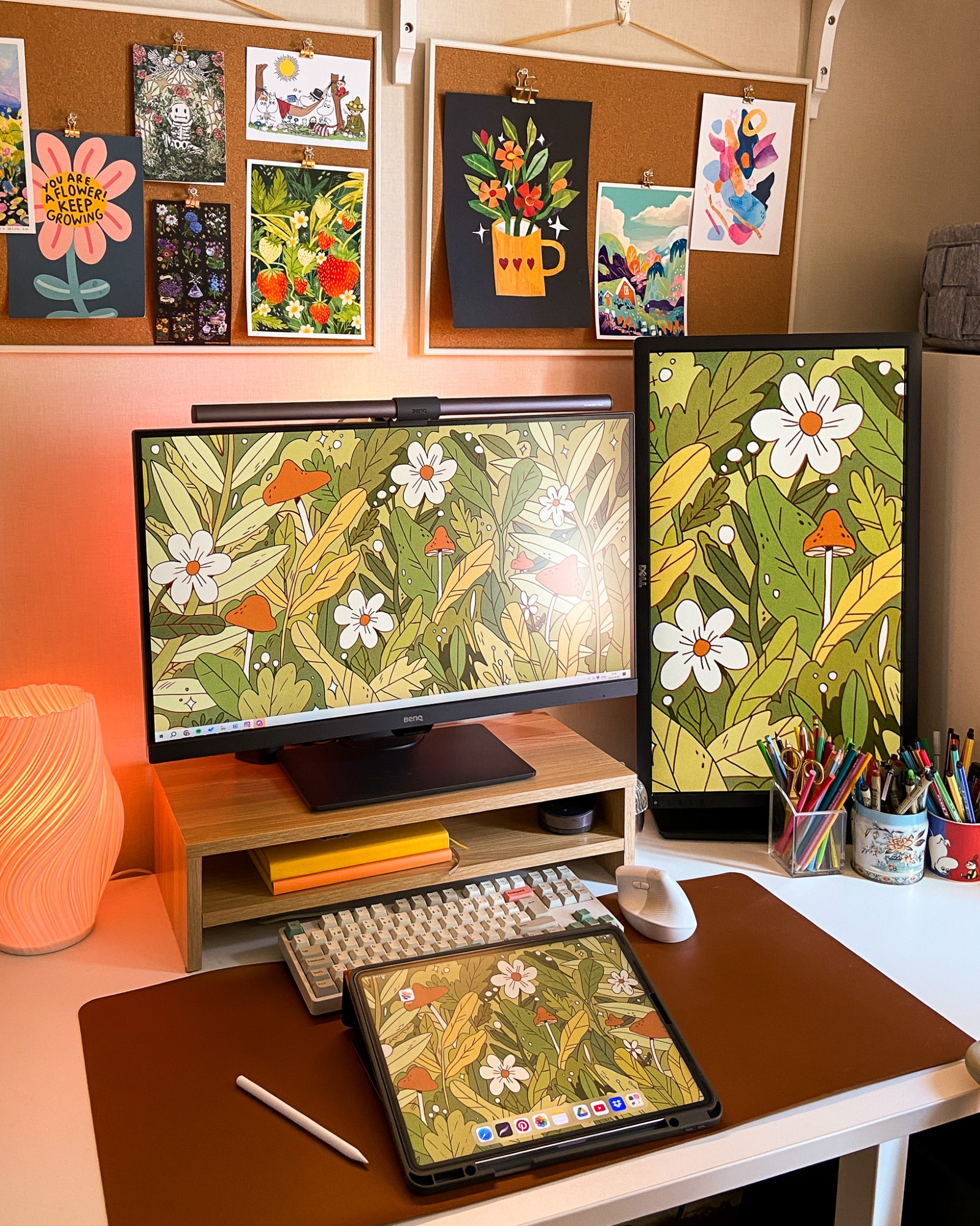 A photo of my desk setup in 2024, lots of warm tones and cozy vibes. There's a brown desk mat, a cute lamp, two monitors and an iPad with the same drawing as wallpaper. Above the monitors are two cork boards with lots of art on them. To the right on the desk are cups with pens and pencils in them. 