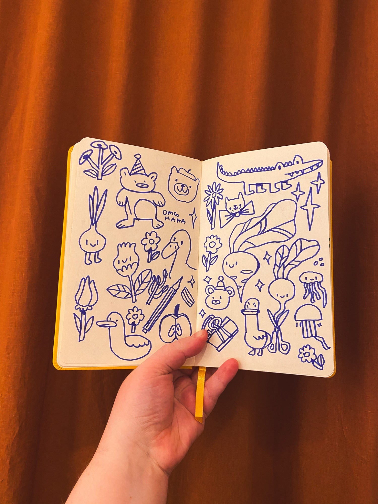 A photo of my hand holding my sketchbook, inside of which are some silly doodles made with a blue marker. There is an onion with legs, a crocodile, a very wonky bear with the text "Omg haha" next to it, a duck, an angry goose, a happy beet, a radish, and lots of flowers. Behind the sketchbook is a rusty brown curtain, and the lighting is soft and warm.