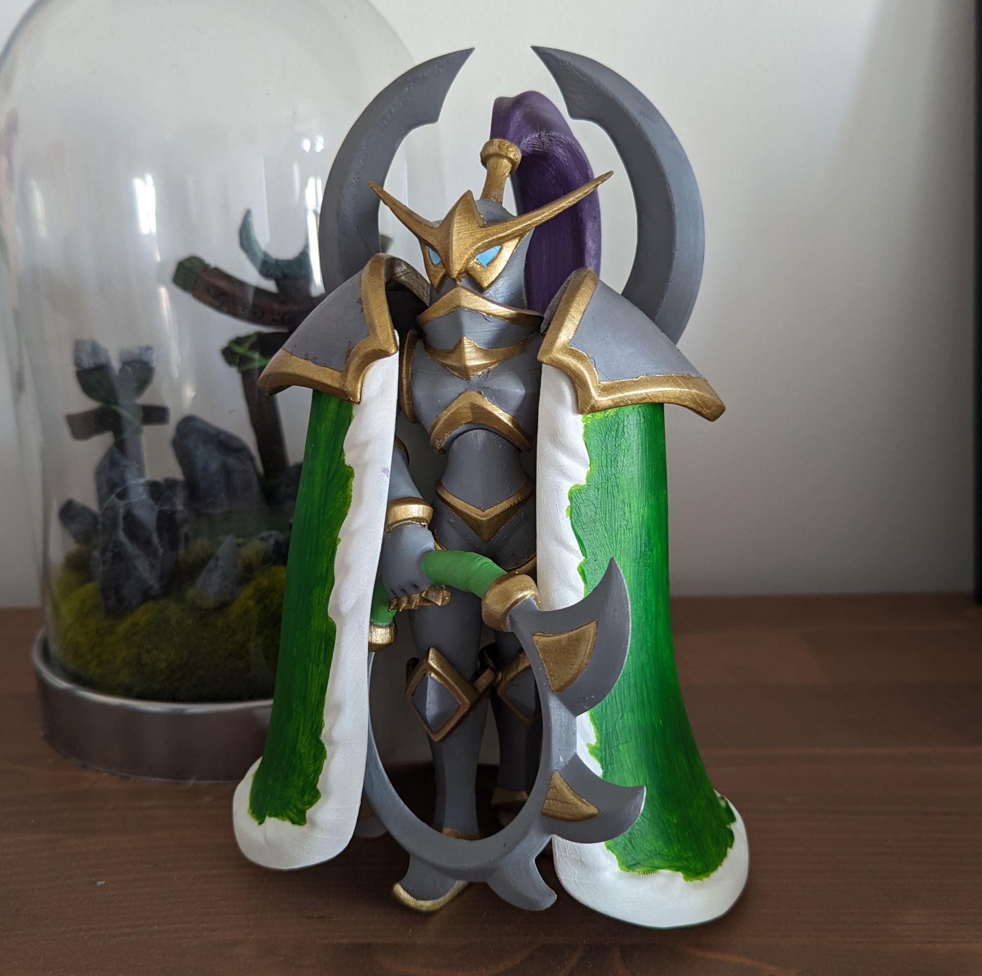 Figurine of the character Maeiv from World of Warcraft placed on dark wood with a white background. The character wears full silver and green armor with an owl-shaped helmet. Maeiv has a ponytail of purple hair that sticks out from her helmet. She also carries a circular, toothed glaive weapon which is partially hidden by her long covering green cape. 
Behind her stands a Moonwell sculpture also from the Warcraft universe in green and stone tones.