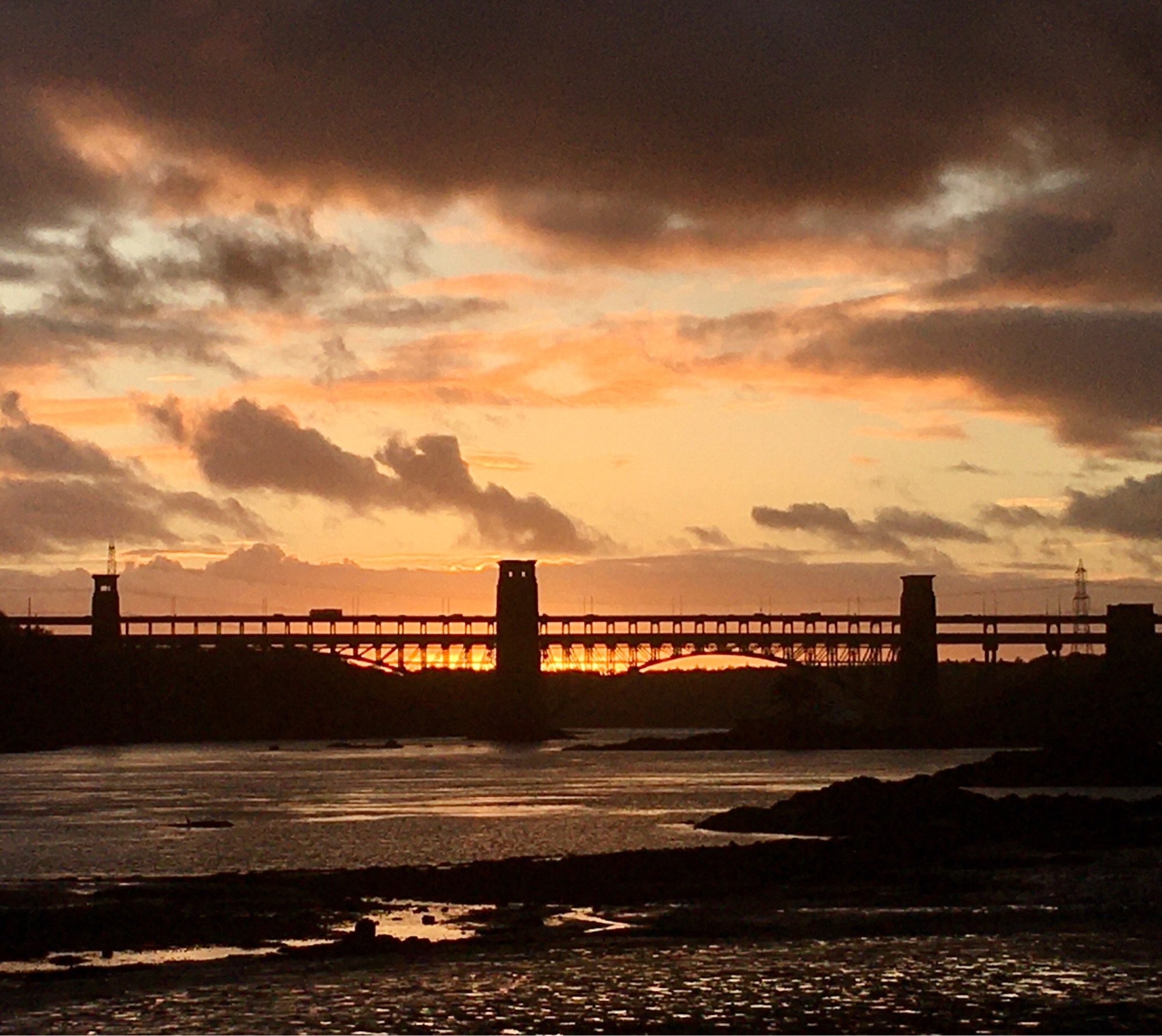 Pale sunset in heavy cloud behind a distant split-level bridge spanning a strait of seawater, over which a few vehicles are crossing on the top deck. The railway level is empty.