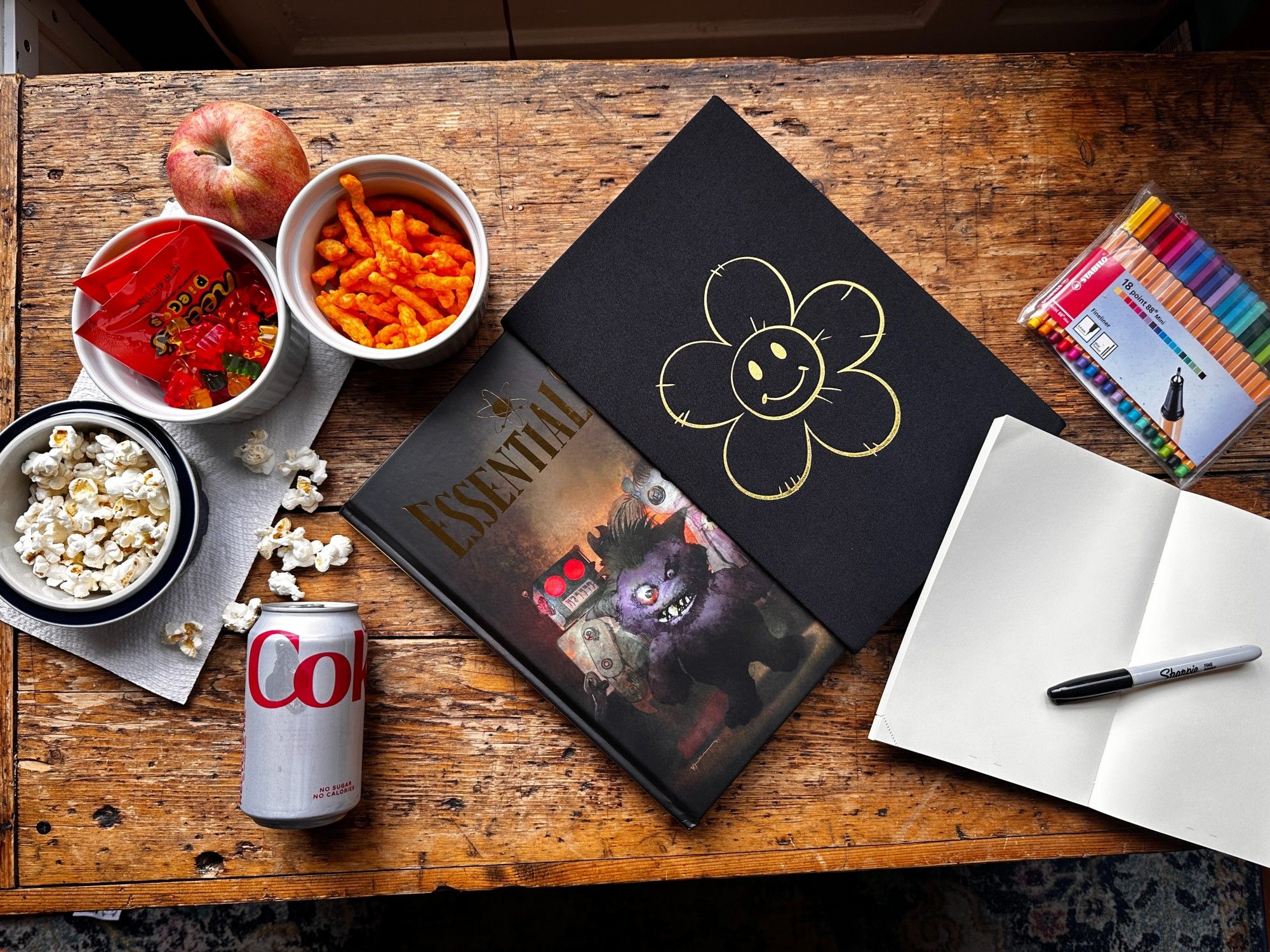 A horizontal photo of ESSENTIALS half way out of its case. It is surrounded by snacks like popcorn and Diet Coke on the left and a blank drawing pad with a sharpie laid on top of it on its right. There is a packet of markers above the drawing pad. All items are on a wood grain background.