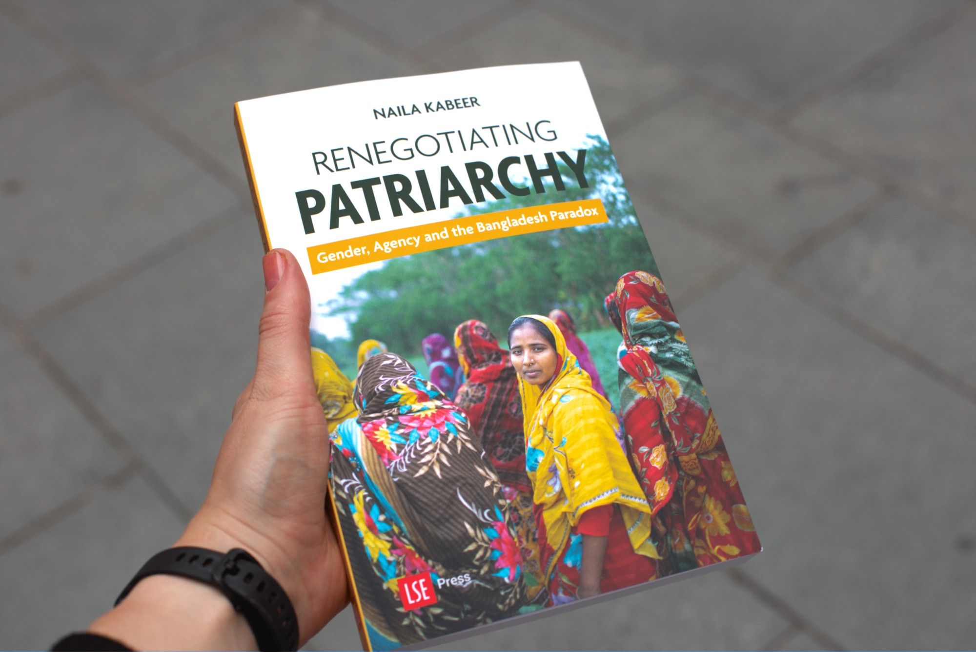 A hand holds a copy of Naila Kabeer's new book, Renegotiating Patriarchy. The cover shows a group of women in colourful saris walking away from the camera. A woman in a yellow sari turns back and looks over her shoulder directly at the camera.