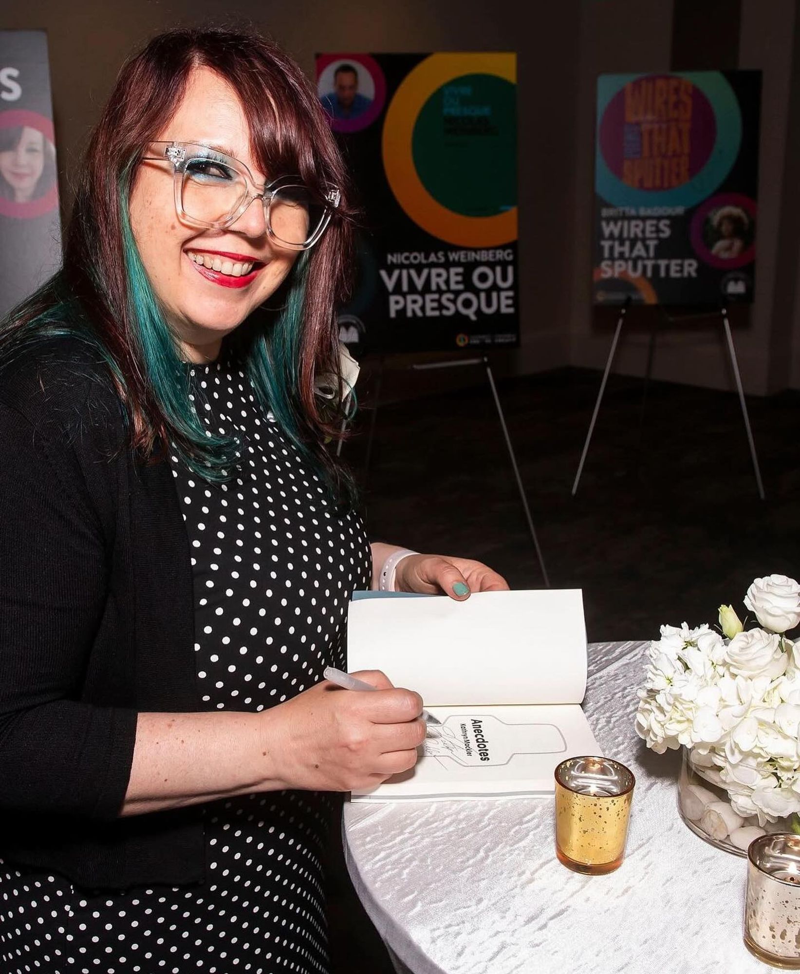 Author Kathryn Mockler, a white woman with red and blue hair, signing a copy of her short story collection, Anecdotes.