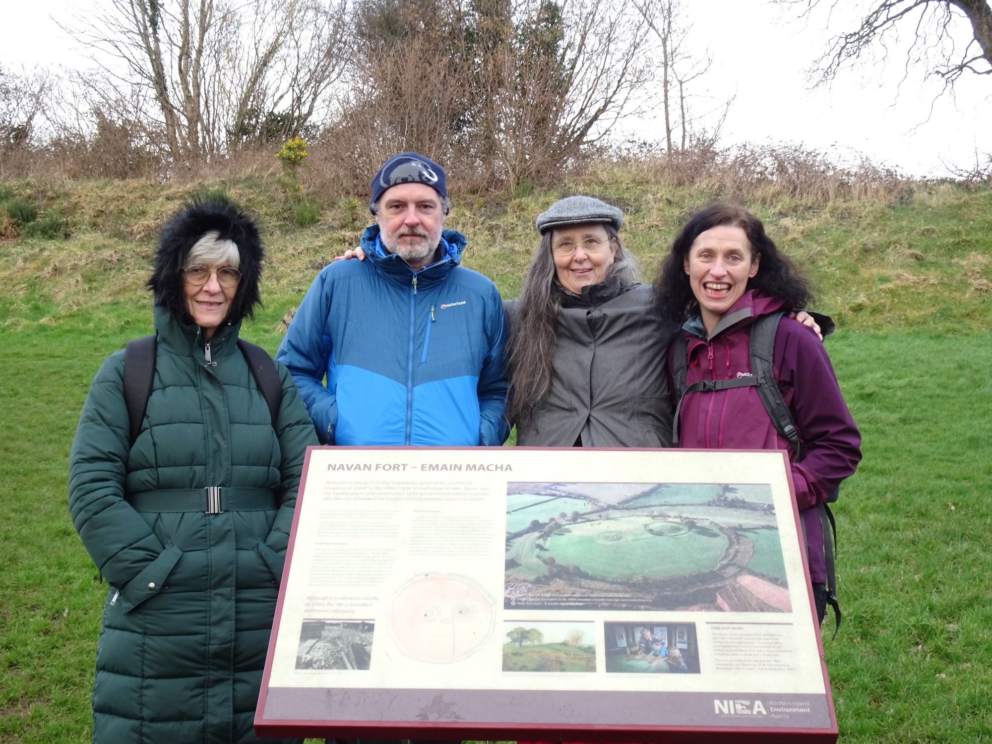 Four gorgeous light skinned humans in front of a sign about Navan Fort - Emain Macha. Leafless trees and scrub in background.