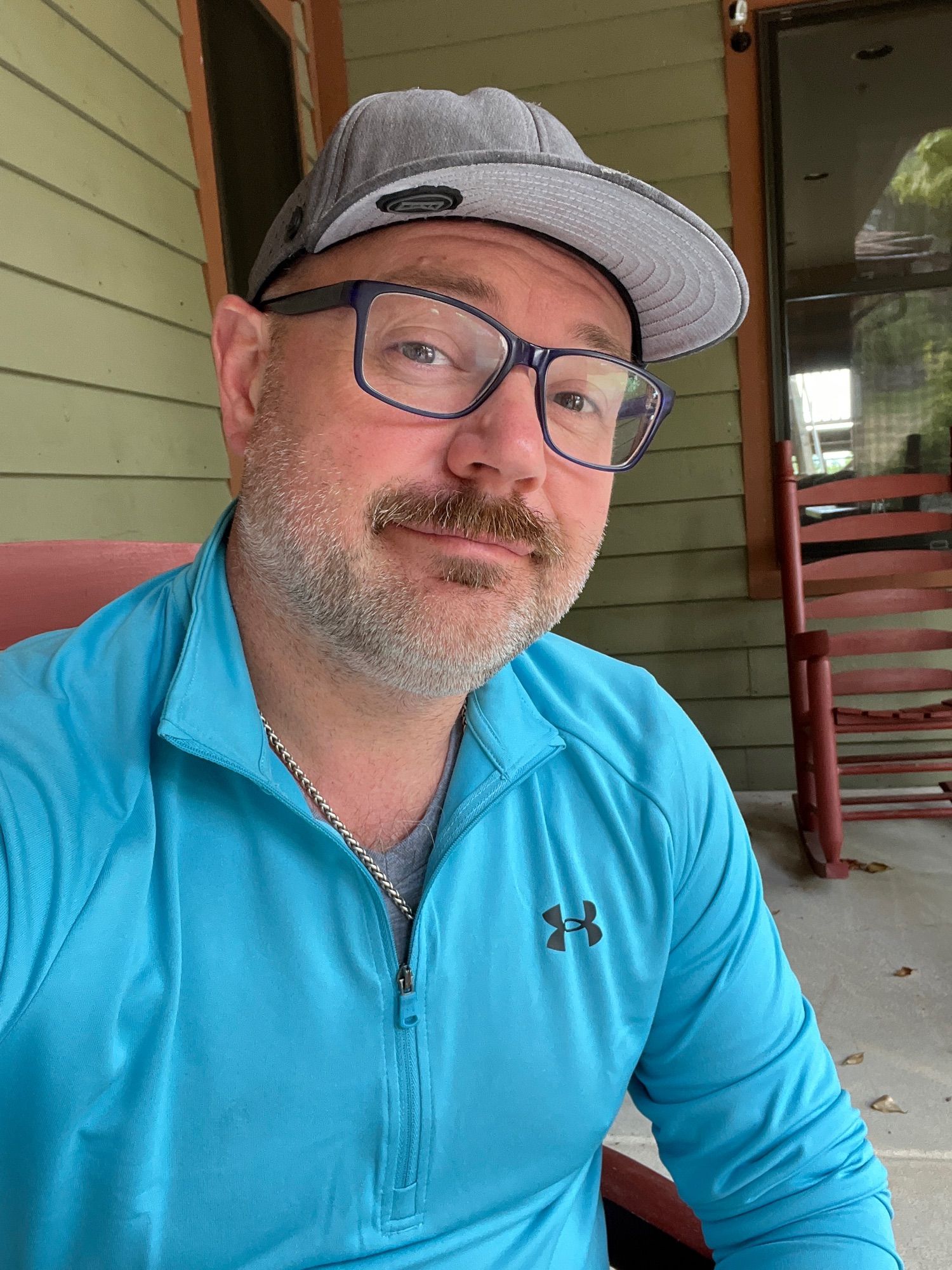 Middle Aged man wearing a hat and glasses, Blue under armor pull over with a mustache sitting on a porch with rockers.