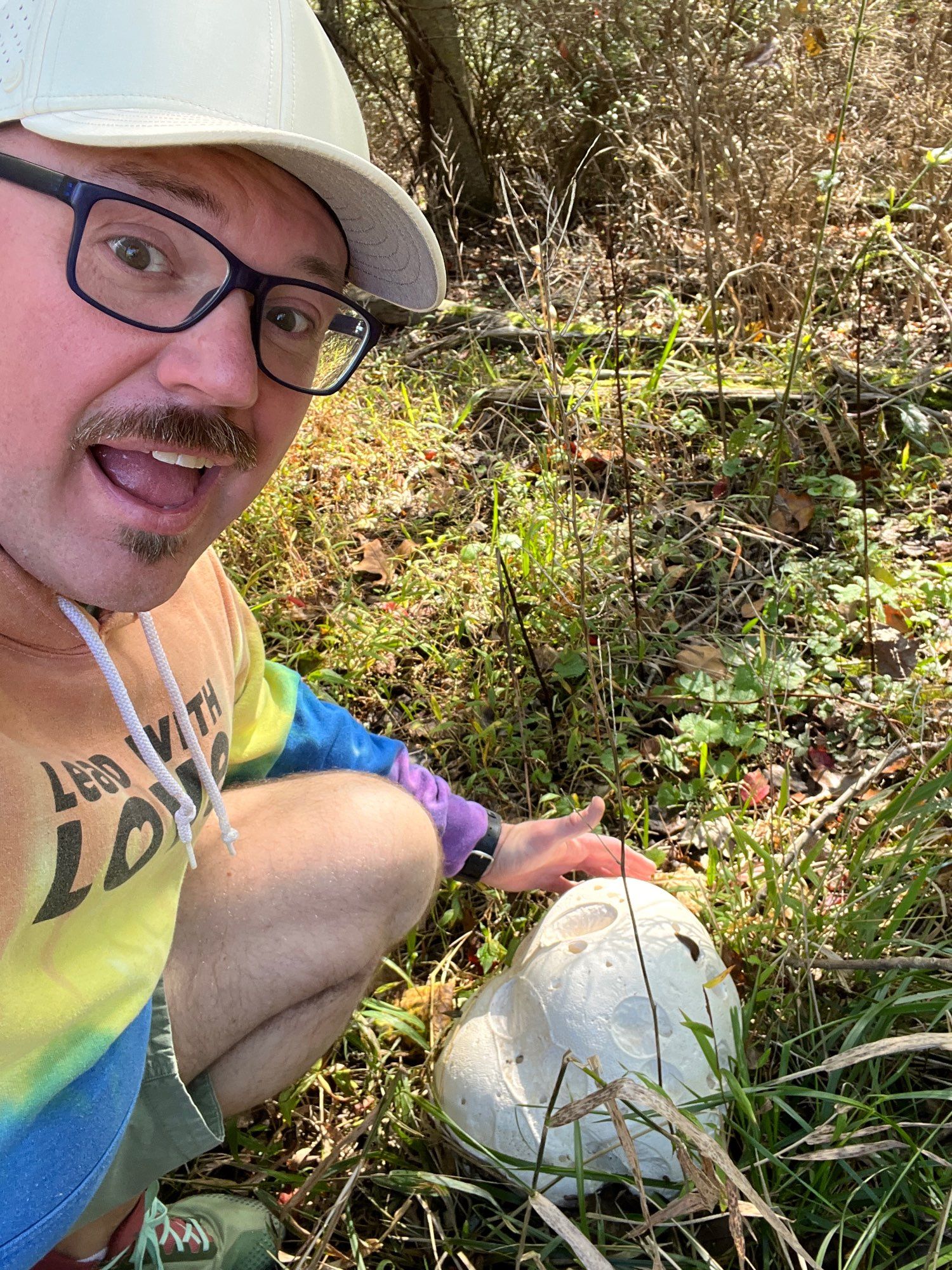 Man with glasses and rainbow tie dyed hoodie finding large puffball mushroom in forested area.