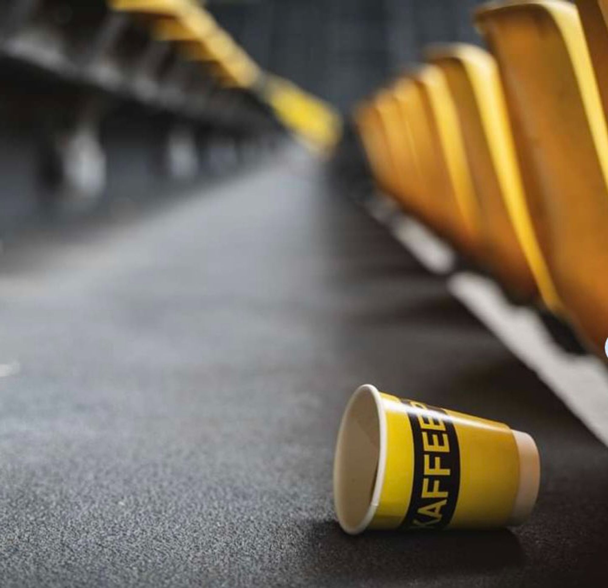 Auf dem Boden der Stadionränge liegt ein Becher in schwarz gelb. Auf dem Becher steht in gelber Schrift vor schwarzem Hintergrund Kaffee.