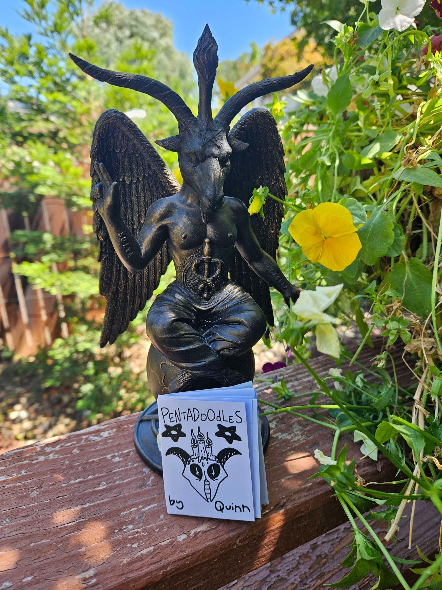 A tiny zine titled, “Pentadoodles by Quinn” rests against a Baphomet statue in my backyard.