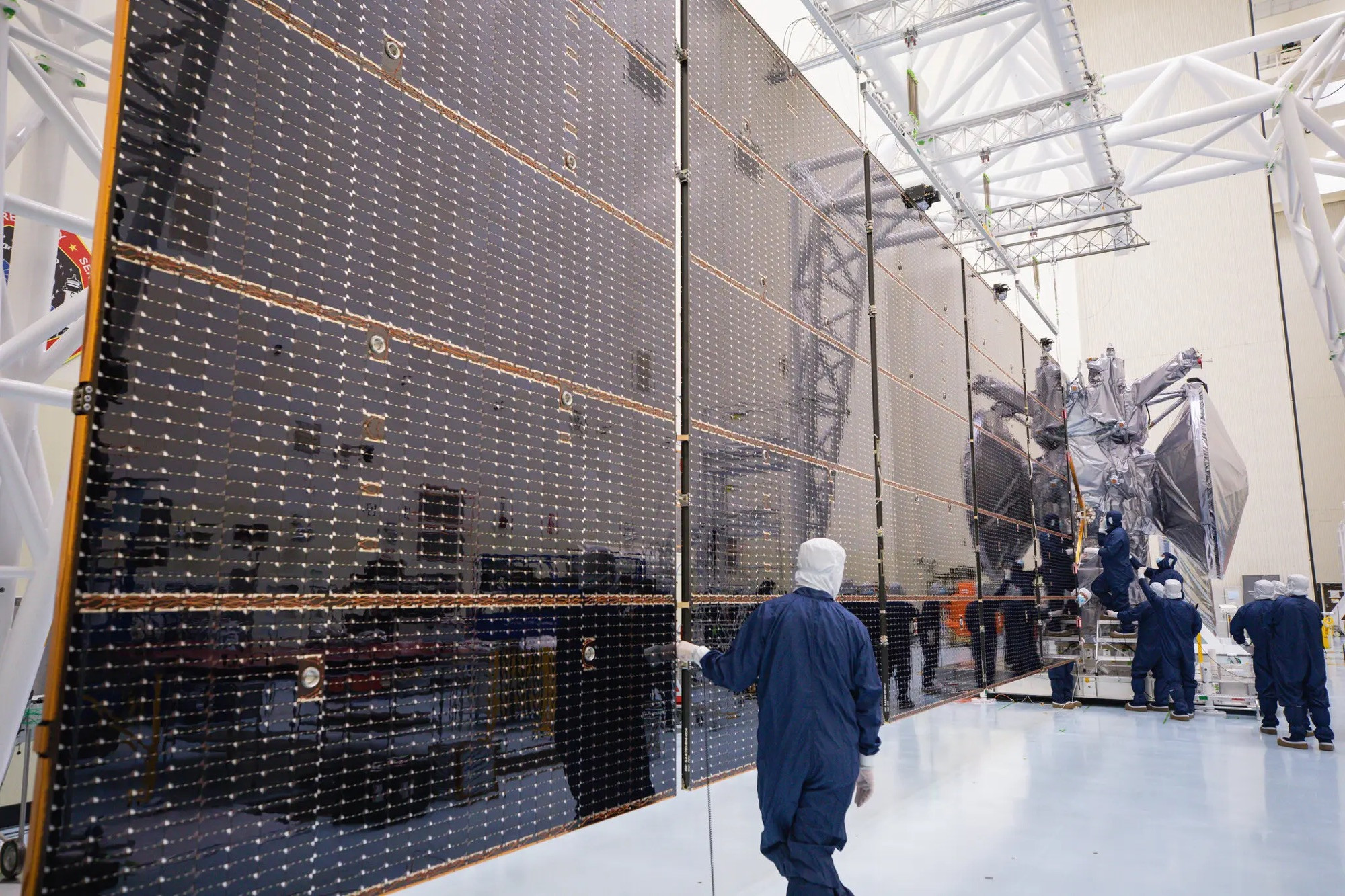 Europa Clipper’s solar arrays are shown with a few people along side. The solar arrays are black with gold lines and are being attached to the spacecraft in the distance in a huge room with white walls and floor. 