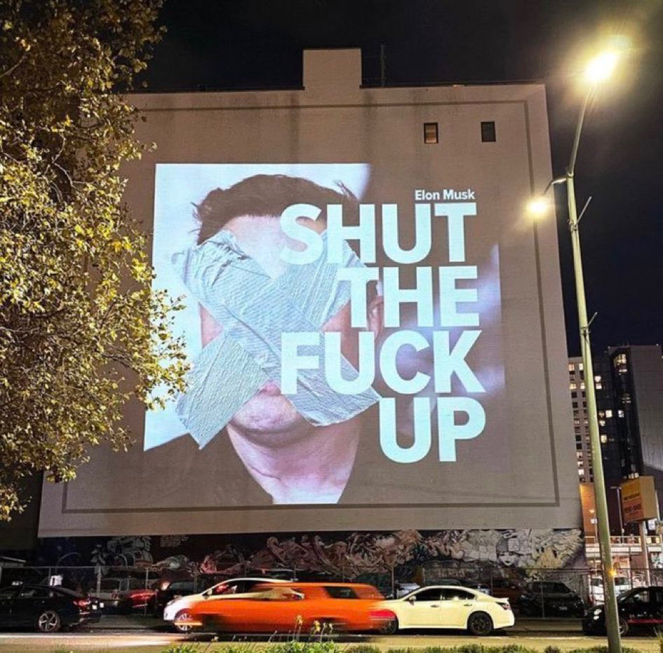 A image on the sign of a building saying shut the fuck up duct taped over Elon musks face