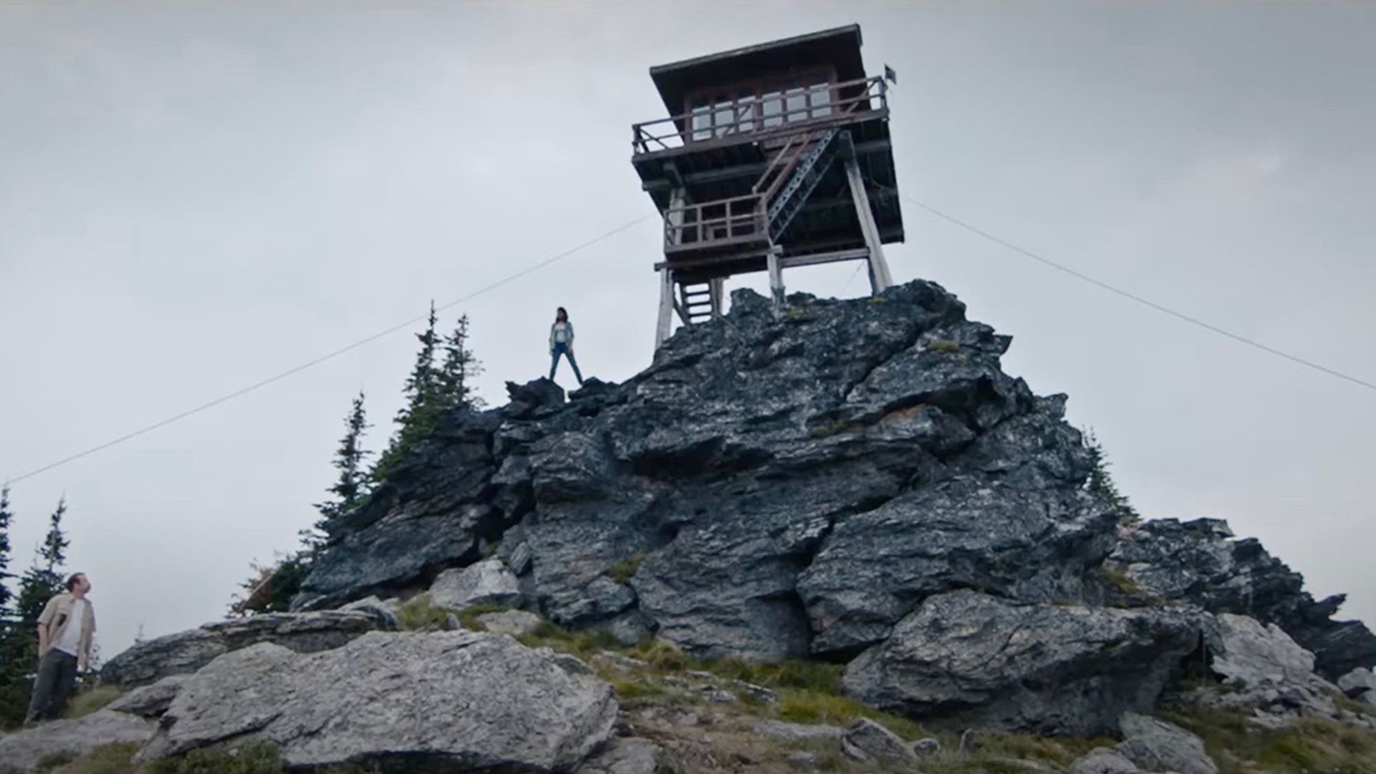 A fire watch outpost sits atop a rocky peak with a woman standing next to the base of it