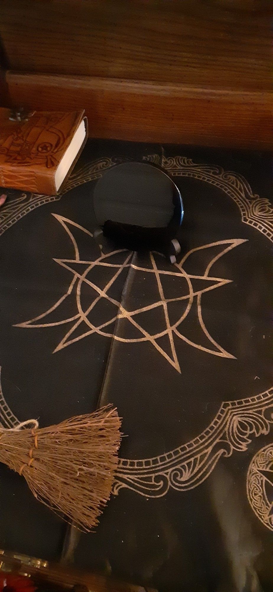 A picture of a black crystal mirror placed upon a cloth with a pentagram/three moon design. There is a mini broom and leather bound book visible at the edges of the image.