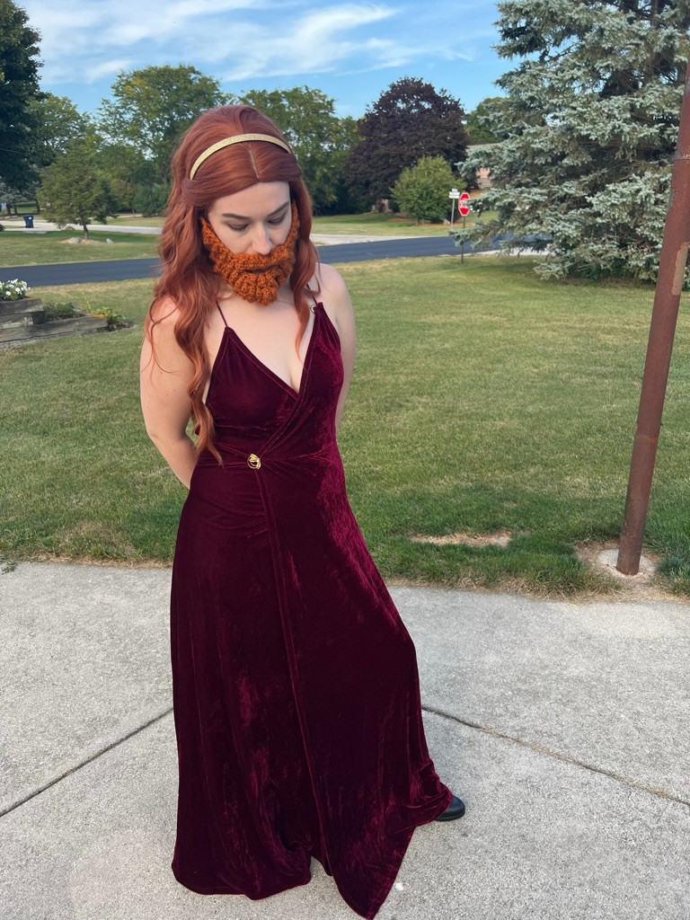 Claire, dressed as Gemma Bronzebeard from NADDPOD, wears a long red wig, maroon dress with gold broach, and red beard. She looks to the ground, outdoors in a suburb on concrete. 