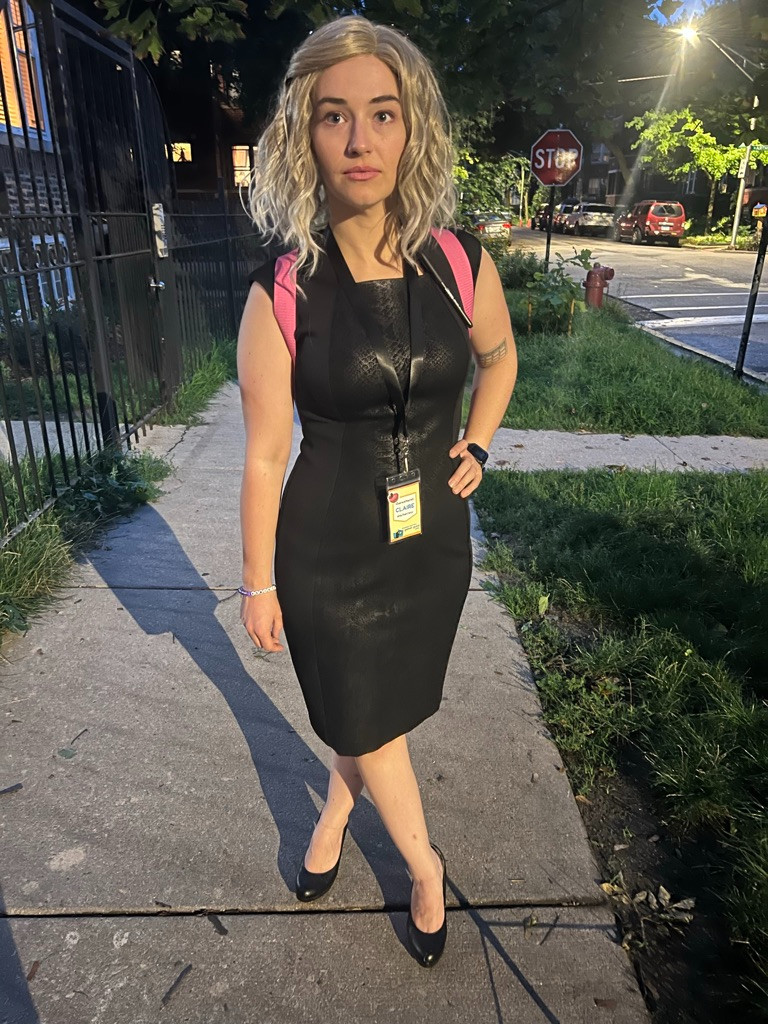 Claire, in a Girlboss Barbie cosplay, looks vapidly and emptily past camera. She wears a Calvin Klein business bitch dress, lanyard, and black heels. She stands on an empty sidewalk with blonde hair and vibrant Barbie-esque makeup. Her hand is on her hip and her leg is crossed in front of her. 