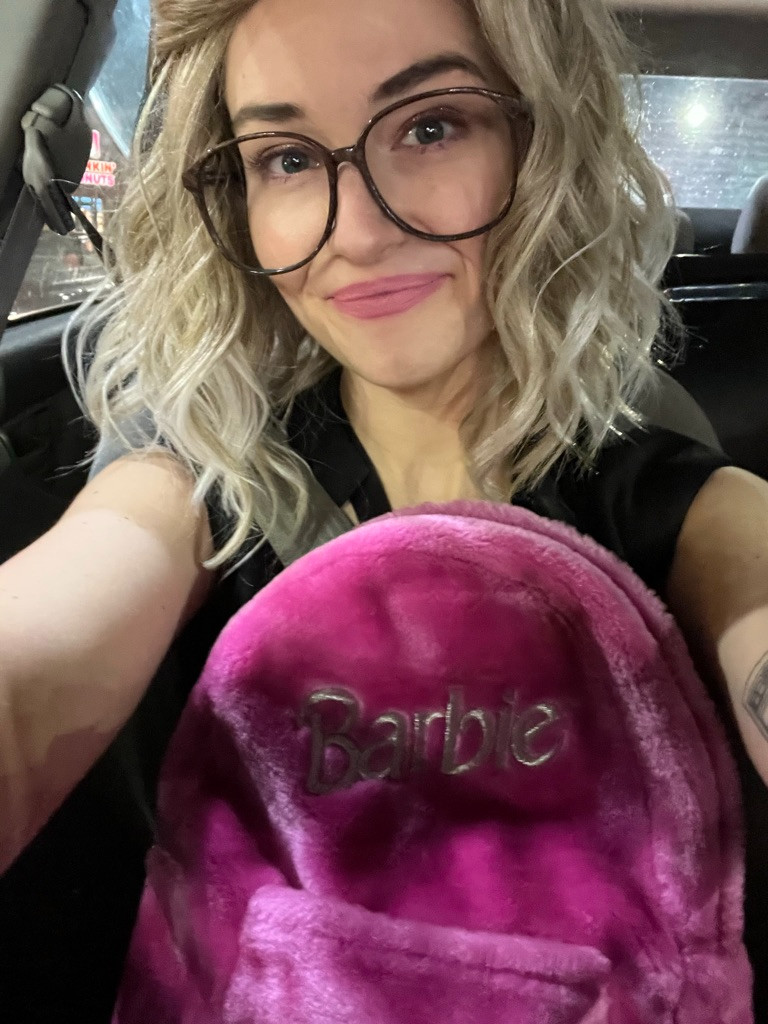 Claire, in a Girlboss Barbie cosplay, has taken a car selfie with large glasses, blonde hair, and vibrant Barbie-esque makeup. A vintage furry Barbie backpack takes up a portion of the frame.