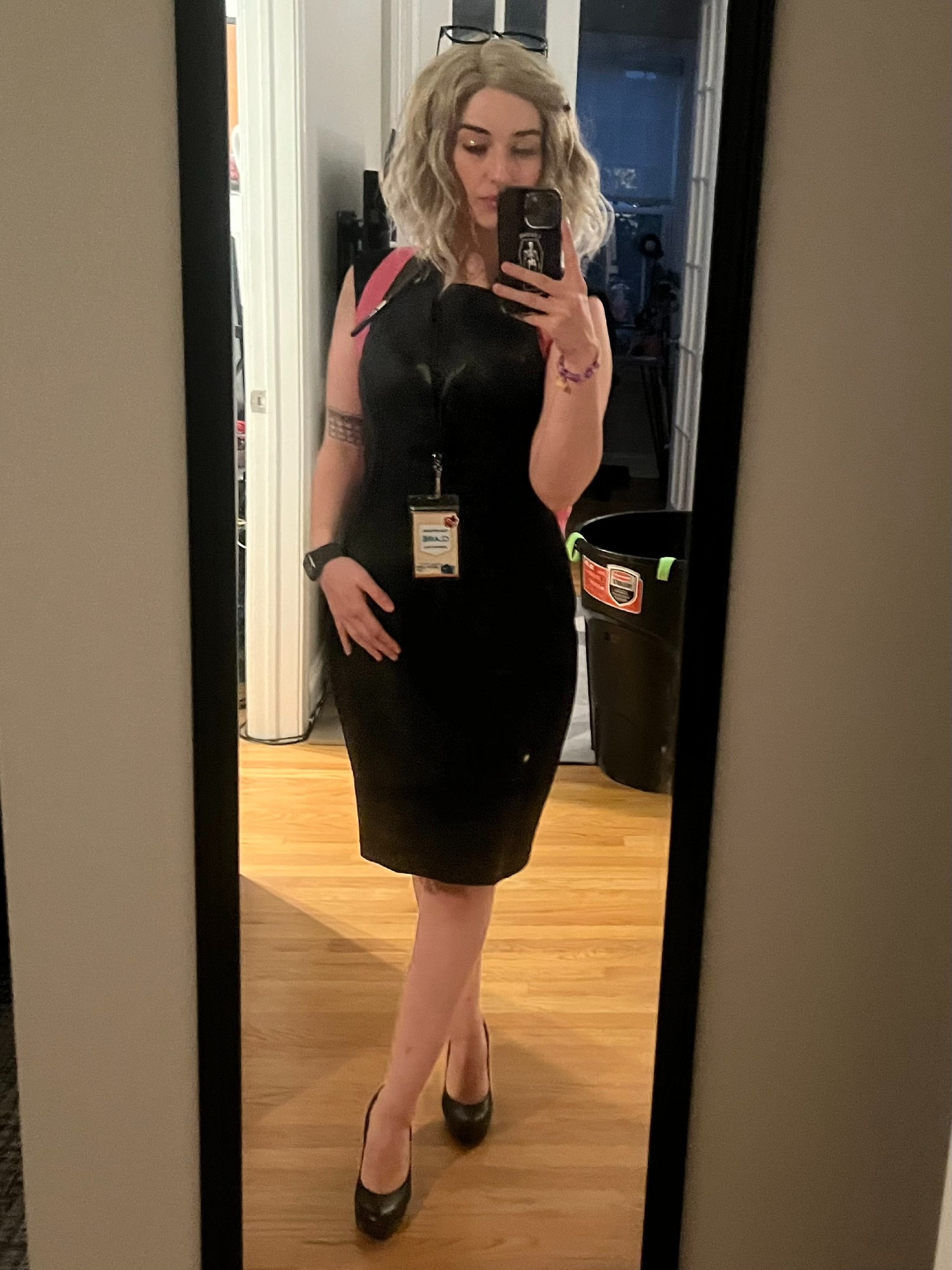 Claire, in a Girlboss Barbie cosplay, takes a mirror selfie. She wears a Calvin Klein business bitch dress, gamer guild lanyard, and black heels. She stands with a hand resting on her hip bone, with blonde hair and vibrant Barbie-esque makeup. She holds her phone and is wearing a pink Barbie backpack. 