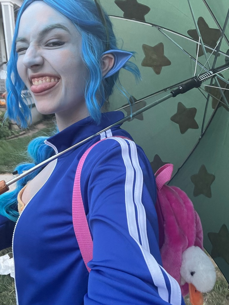 Claire, in a Calliope Petrichor Cosplay, painted light blue with blue ears and a star umbrella, winks at the camera with her tongue slightly out in a selfie. You can see the stuffed duck representing Foster peeking out of her backpack.  