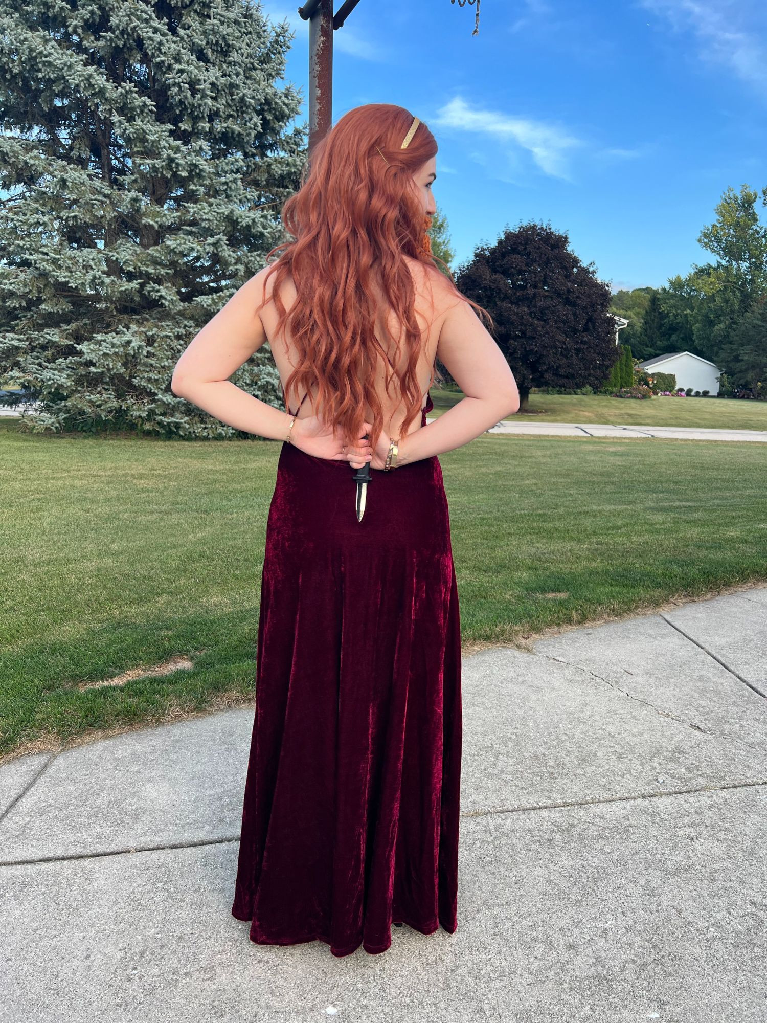 Claire, dressed as Gemma Bronzebeard from NADDPOD, wears a long red wig, maroon dress with gold broach, and red beard. She faces away from camera, holding a fake dagger behind her back. She is outdoors in a suburb on concrete. 