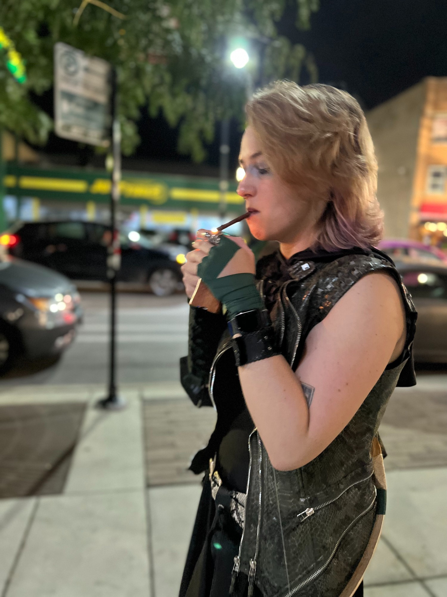 Claire, dressed as Egwene Kindleaf from Not Another D&D Podcast, stands outside, lighting a wooden cigarette. The city street is blurry behind her and you can see a bow over her back. 