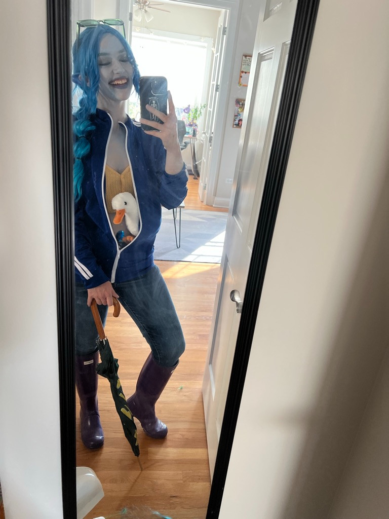 Claire, in a Calliope Petrichor Cosplay, painted light blue with blue ears and a star umbrella that is folded closed in a mirror selfie. The duck representing Foster is tucked into her blue track jacket. Her belly button gem is visible and she wears purple rain boots and a smile. 