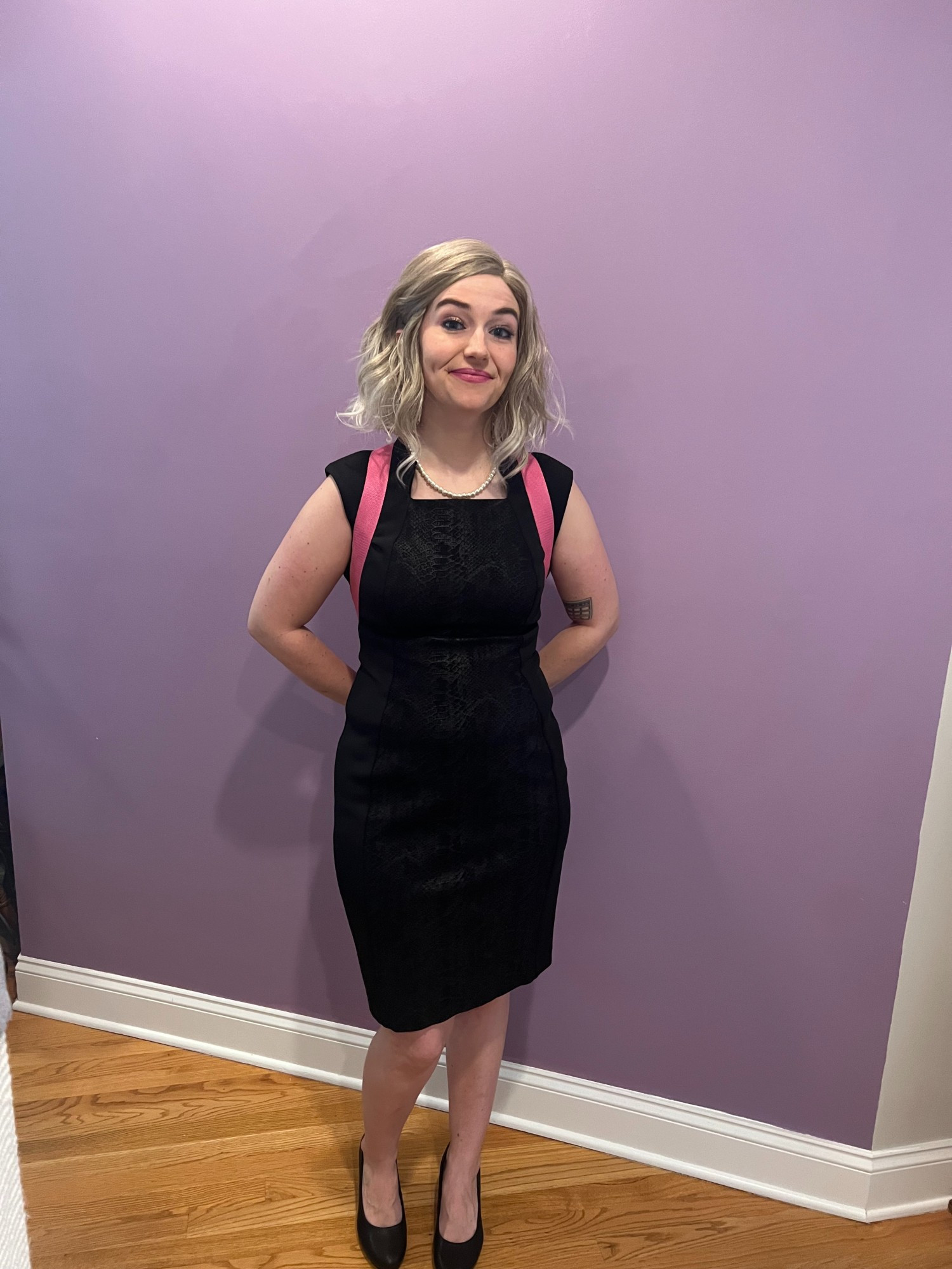 Claire, in a Girlboss Barbie cosplay, wears a Calvin Klein business bitch dress, pearl necklace, and black heels. She stands in front of a purple wall with blonde hair and vibrant Barbie-esque makeup.