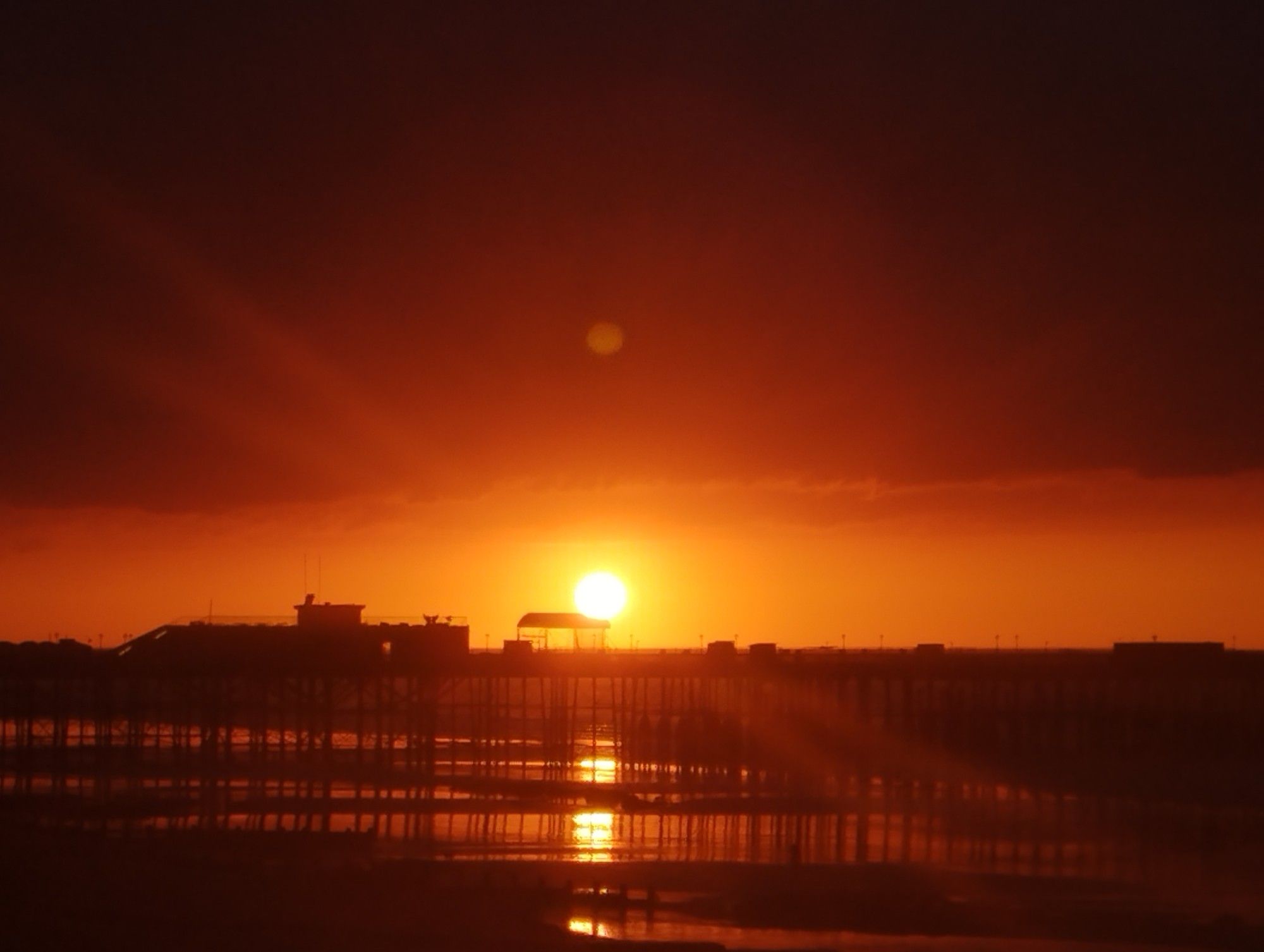 rising sun over Hastings Pier, the sun is a bright orange, between the horizon and heavy overhead cloud cover, everything is deep shades of dark red