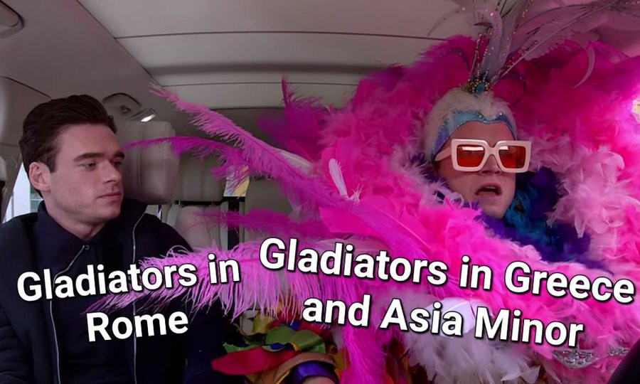 A perplexed man, dressed soberly in black, marked "Gladiators in rome" looks at the man beside him (taron Egerton as elton john) dressed in a massive  pink feather boa, marked "Gladiators in Greece and Asia minor"