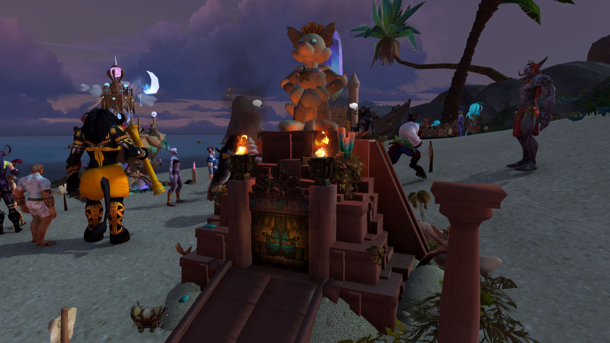 A Warcraft screenshot of a sand castle built in the shape of the pyramid of Atul'amun from Vol'dun, with a sand sculpture of a vulpera with gems for eyes atop it. Its sunset at the beach, partly cloudy, and there's various beachgoers milling about in the background.