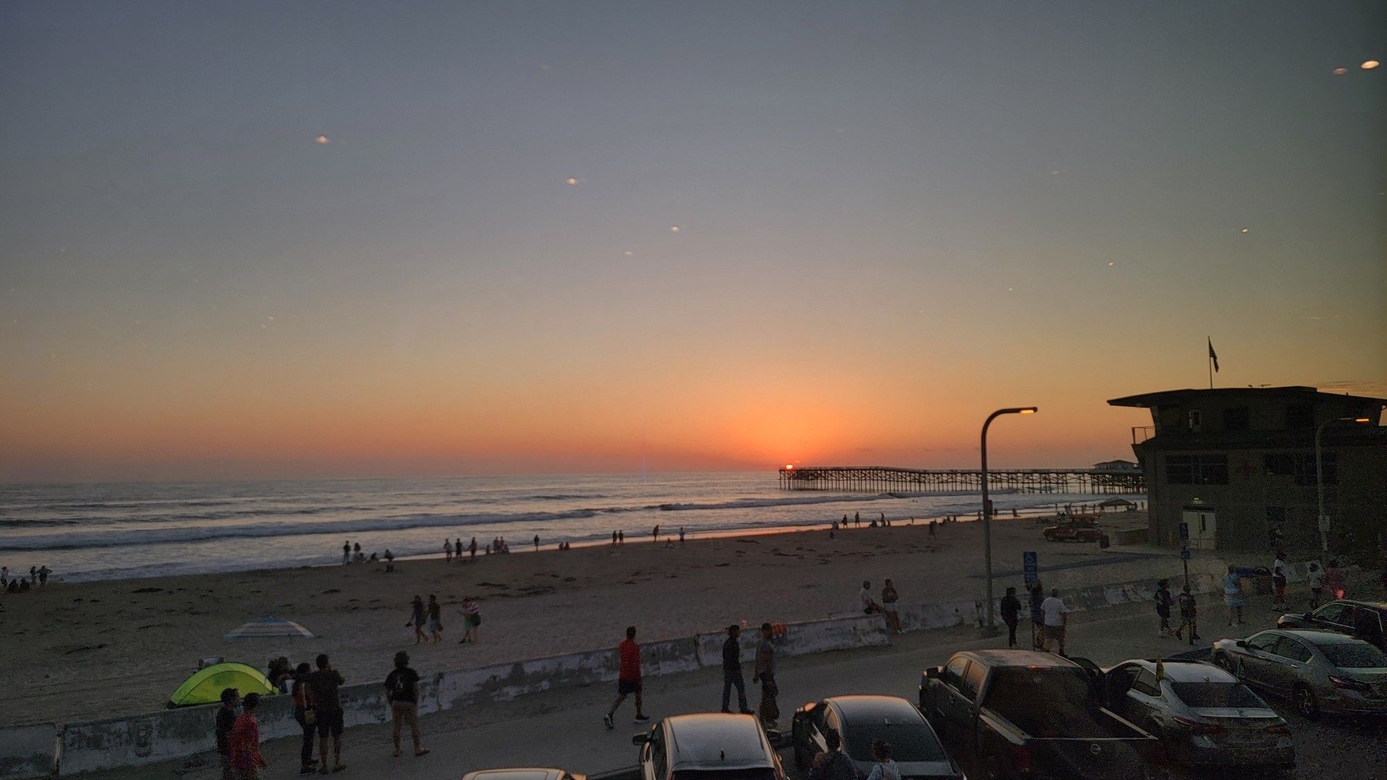 A photo of a beach at late sunset over the water, with people clustered at the water and in the beach sidewalk