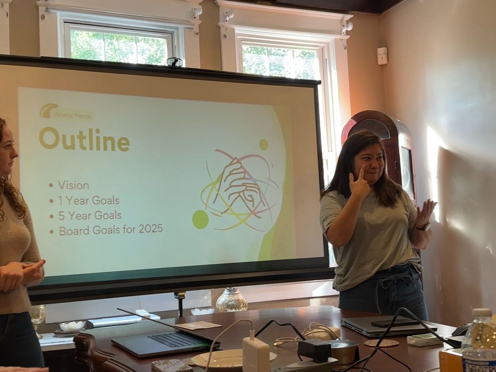 Two women standing in front of a projector screen with “outline: vision, 1 year goals, 5 year goals, board goals for 2025” written in the slide shown