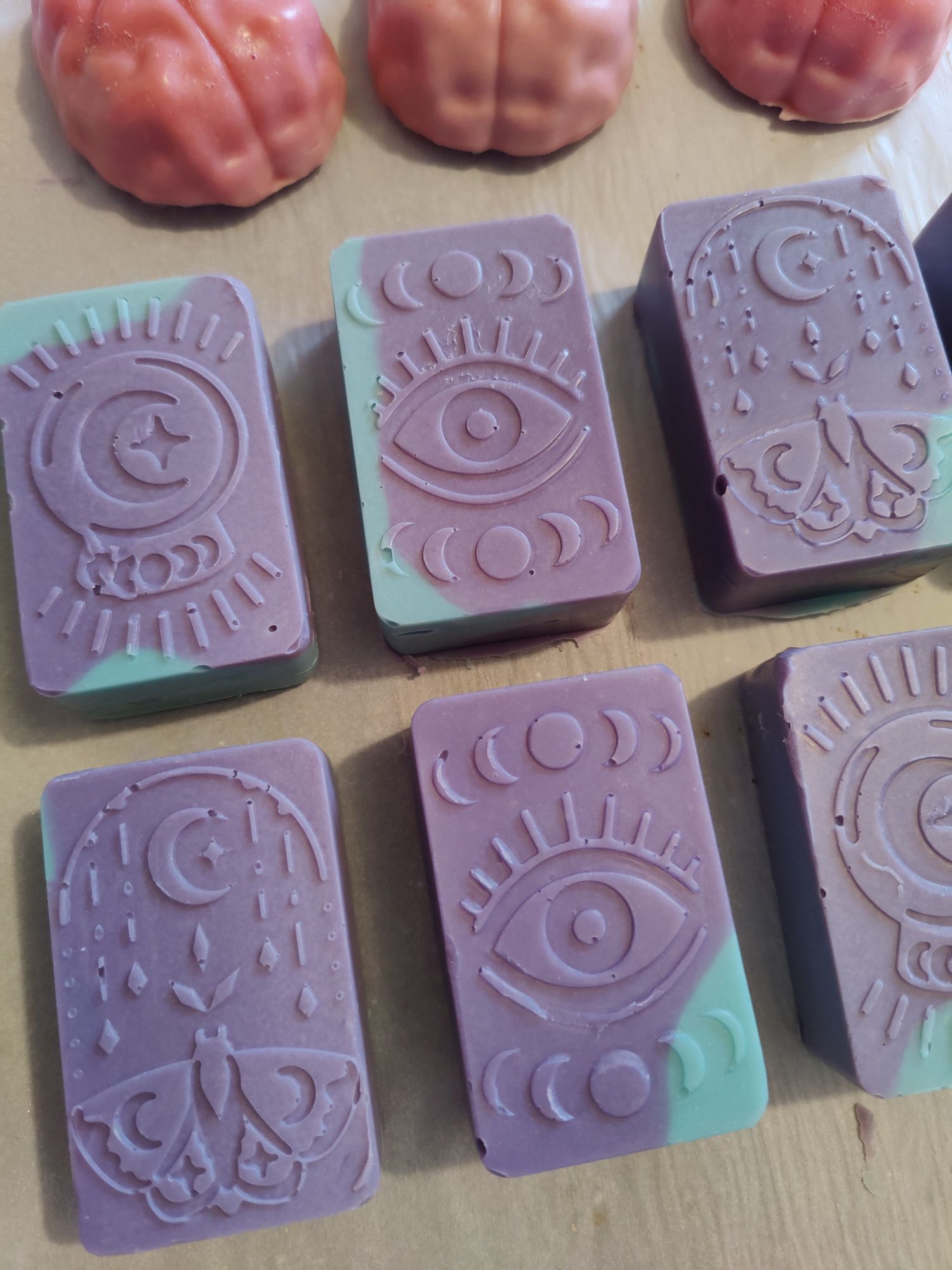 Purple and aqua colored soaps in three different witchy designs. One is an eye with the moon phases above and below. Another is a moth with a crescent moon and crystals. The other is a crystal ball design with a crescent moon and moon phases