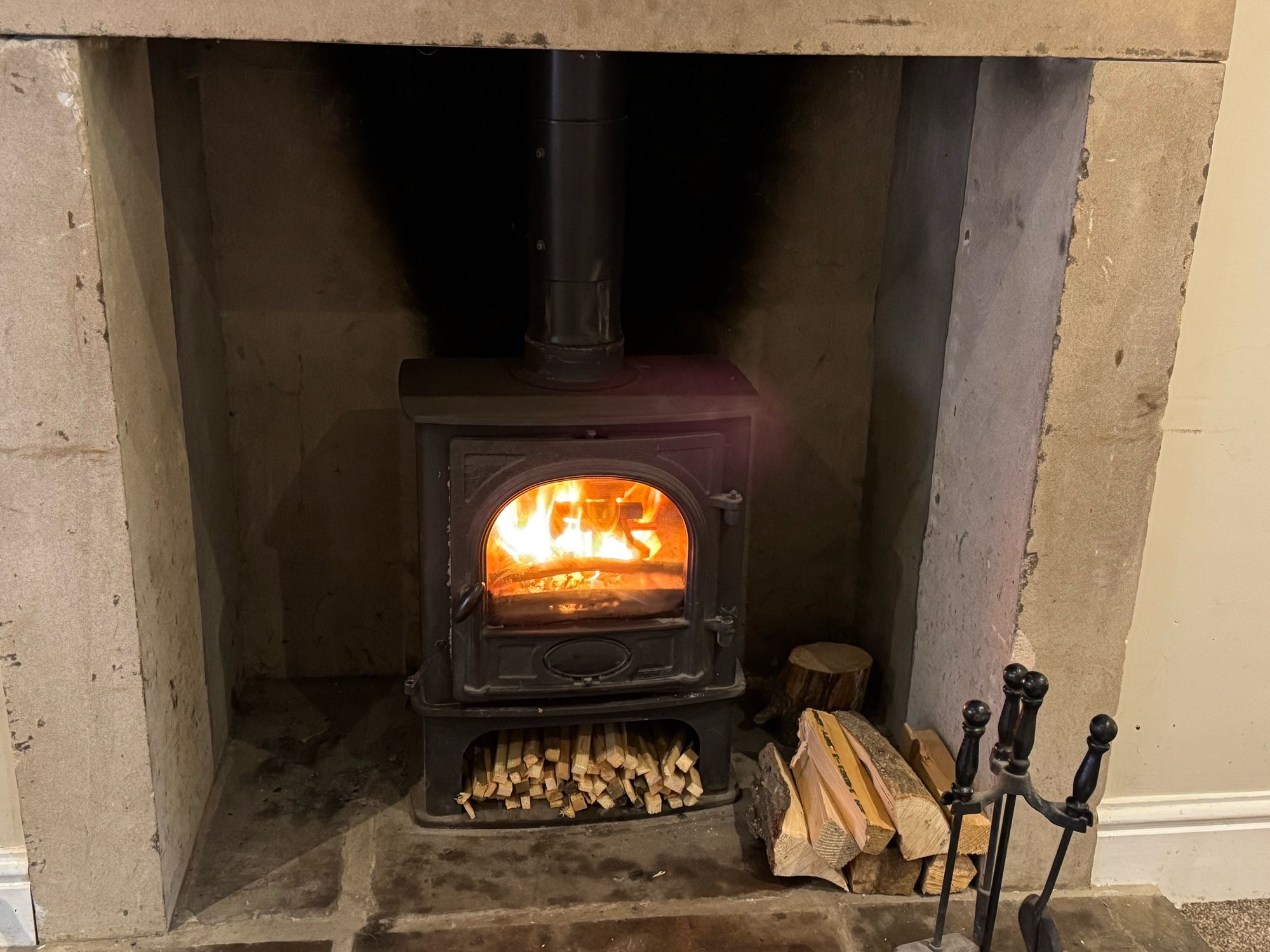 Stone fireplace with a black wood burning stove sitting atop large stone hearth. The fire gas just been lit and is roaring. There is a small pile of logs to the right of the fire place and a poker and shovel set on the hearth