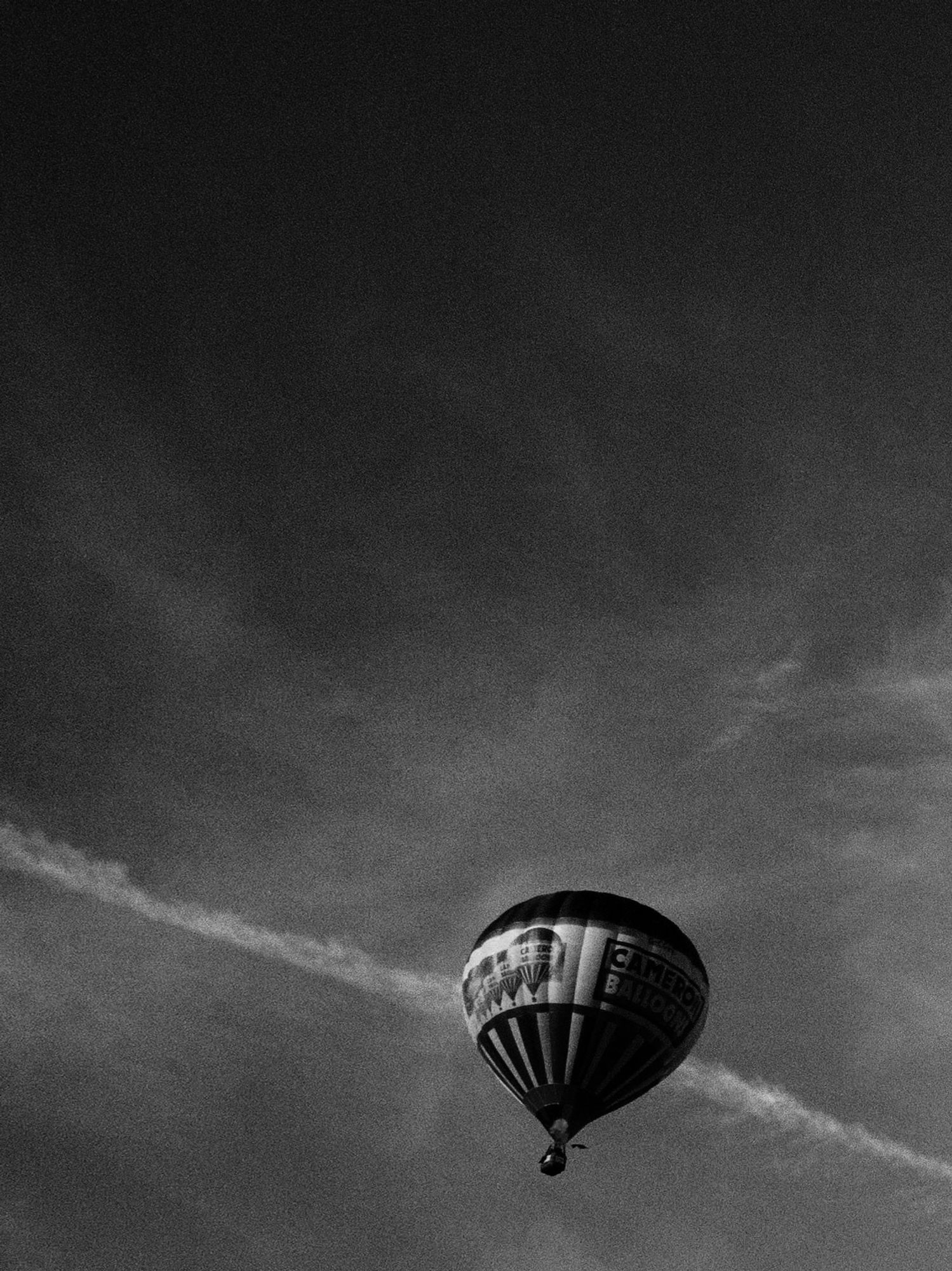 A black and white photo of a hot air balloon with wispy clouds and a plane trail in the background